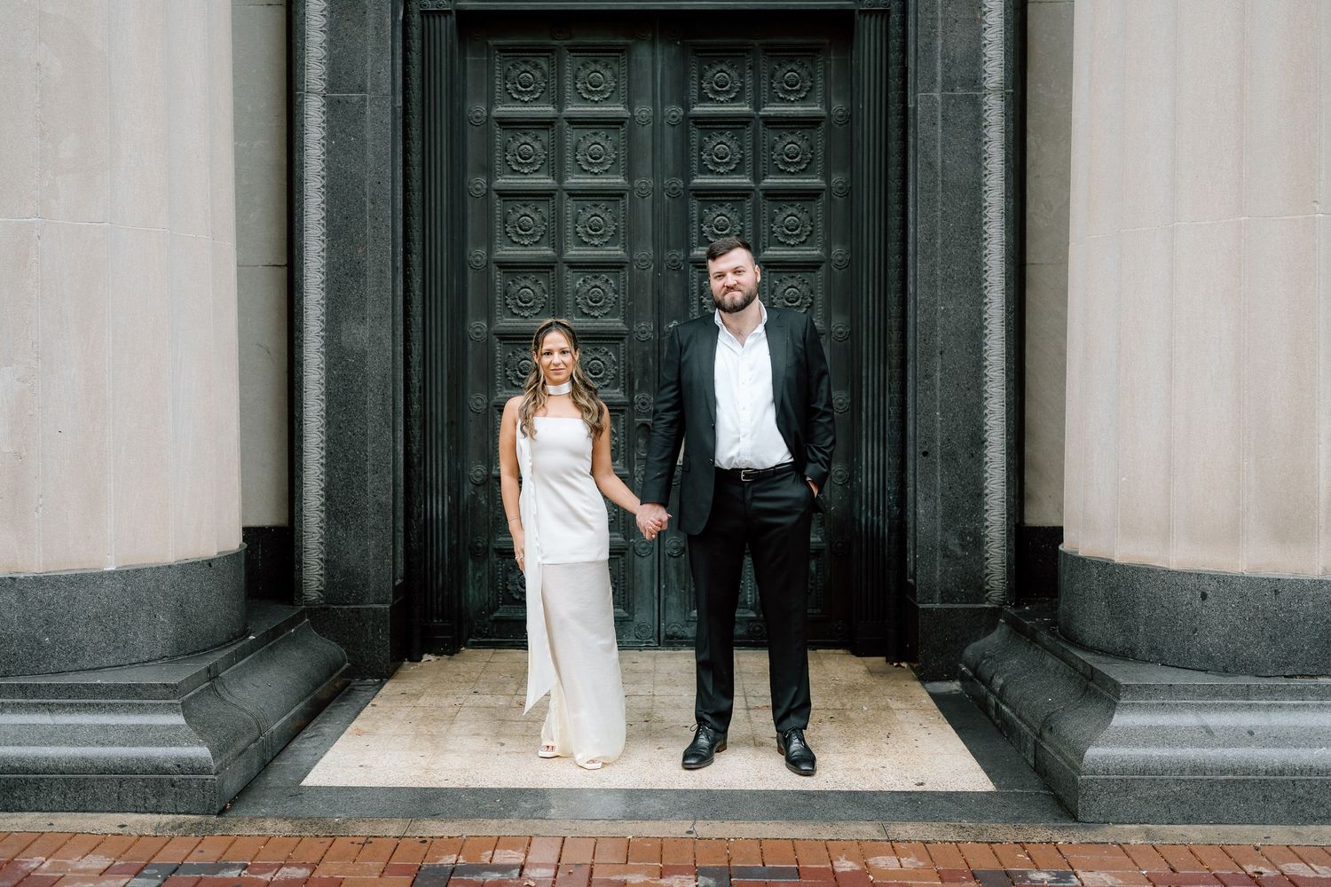 Elegant couple poses in formal attire against ornate dark doors.