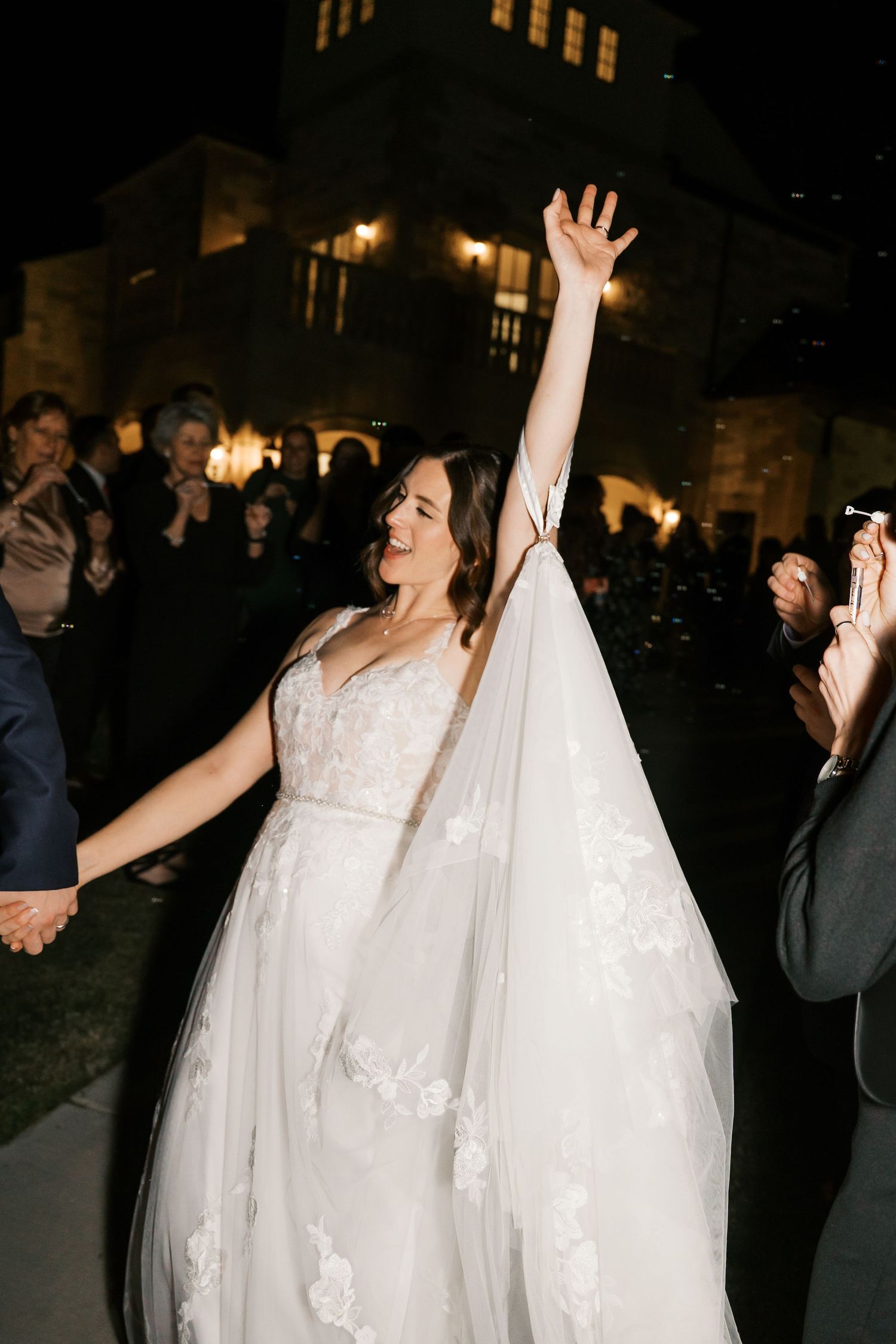 Bride raises her arms in joy during outdoor nighttime wedding celebrations.