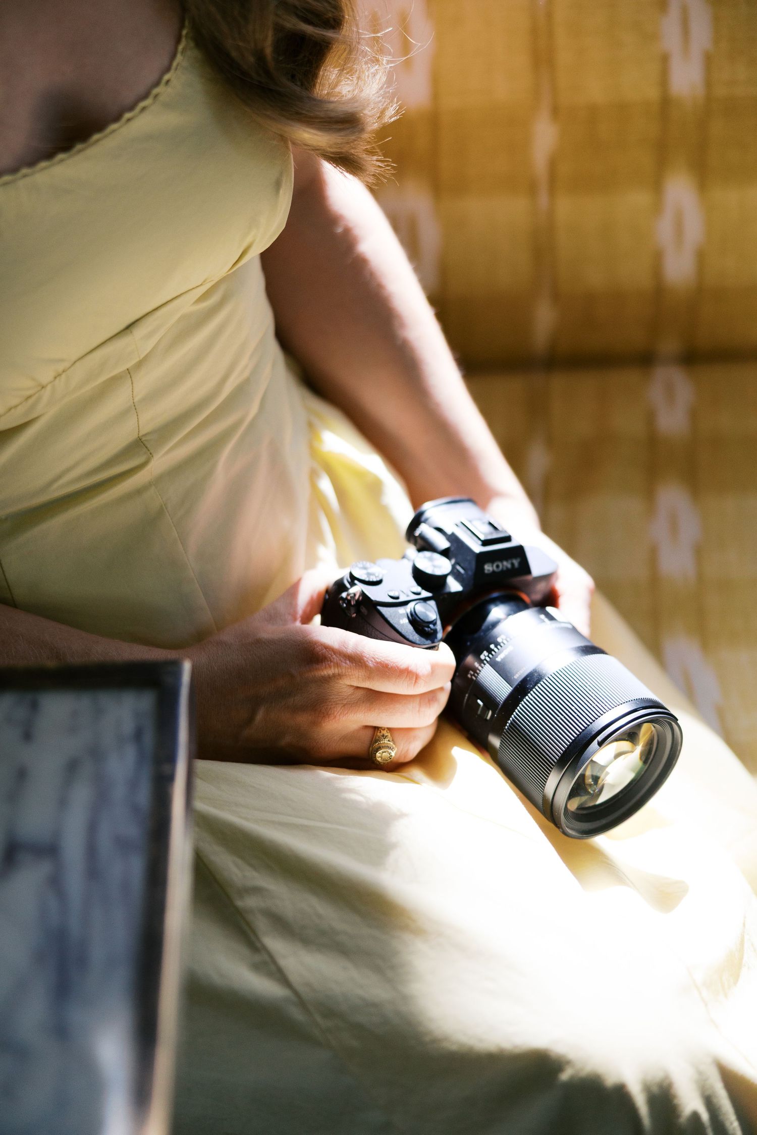 Woman in yellow dress holding a Sony mirrorless camera with zoom lens, seated near a bamboo curtain.
