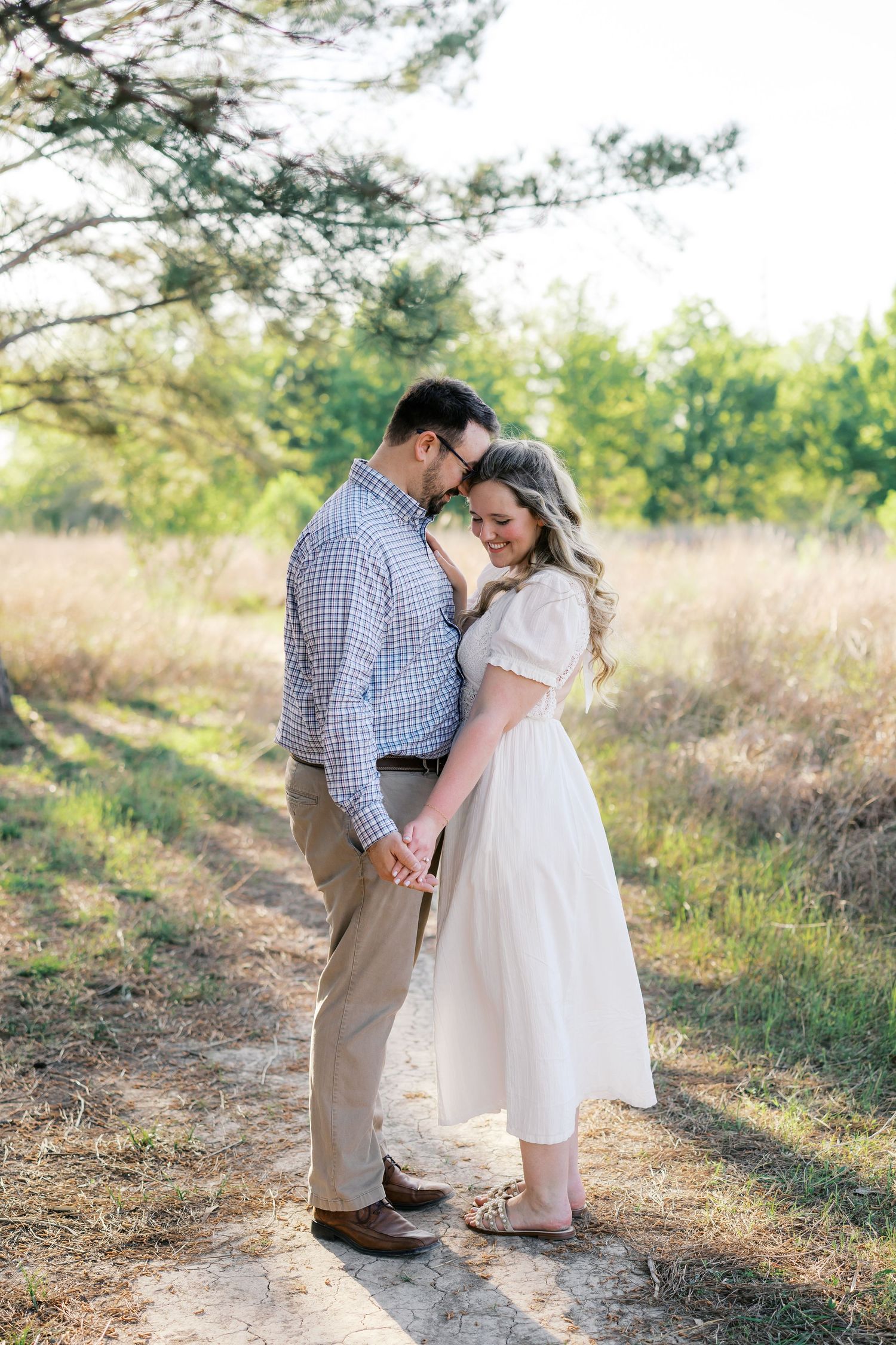 Couple holding hands on a sunlit path in a field, man in plaid shirt and woman in white midi dress, sharing a tender moment.