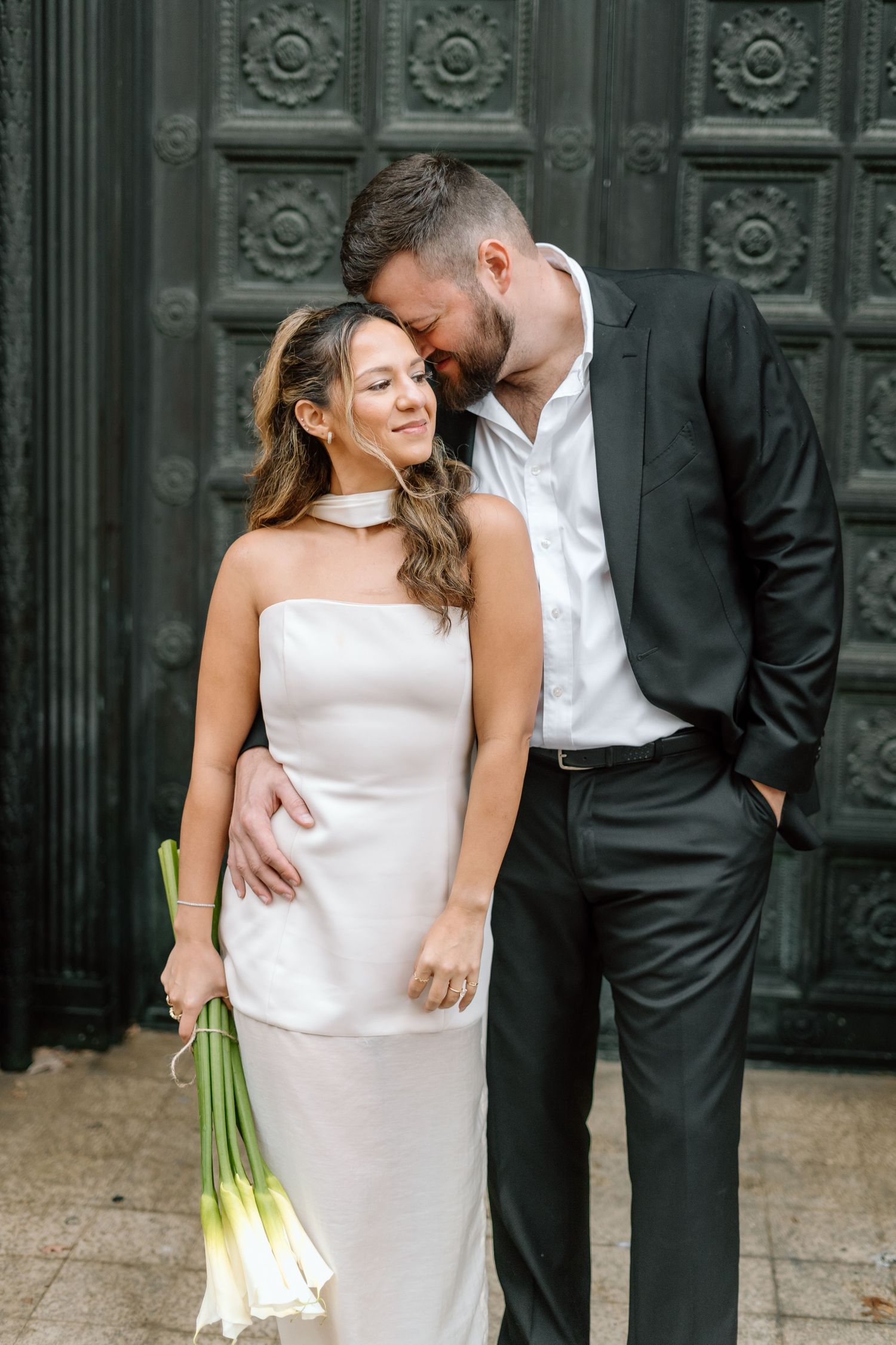 Bride in sleek white dress holds bouquet while posing with groom against dark backdrop.
