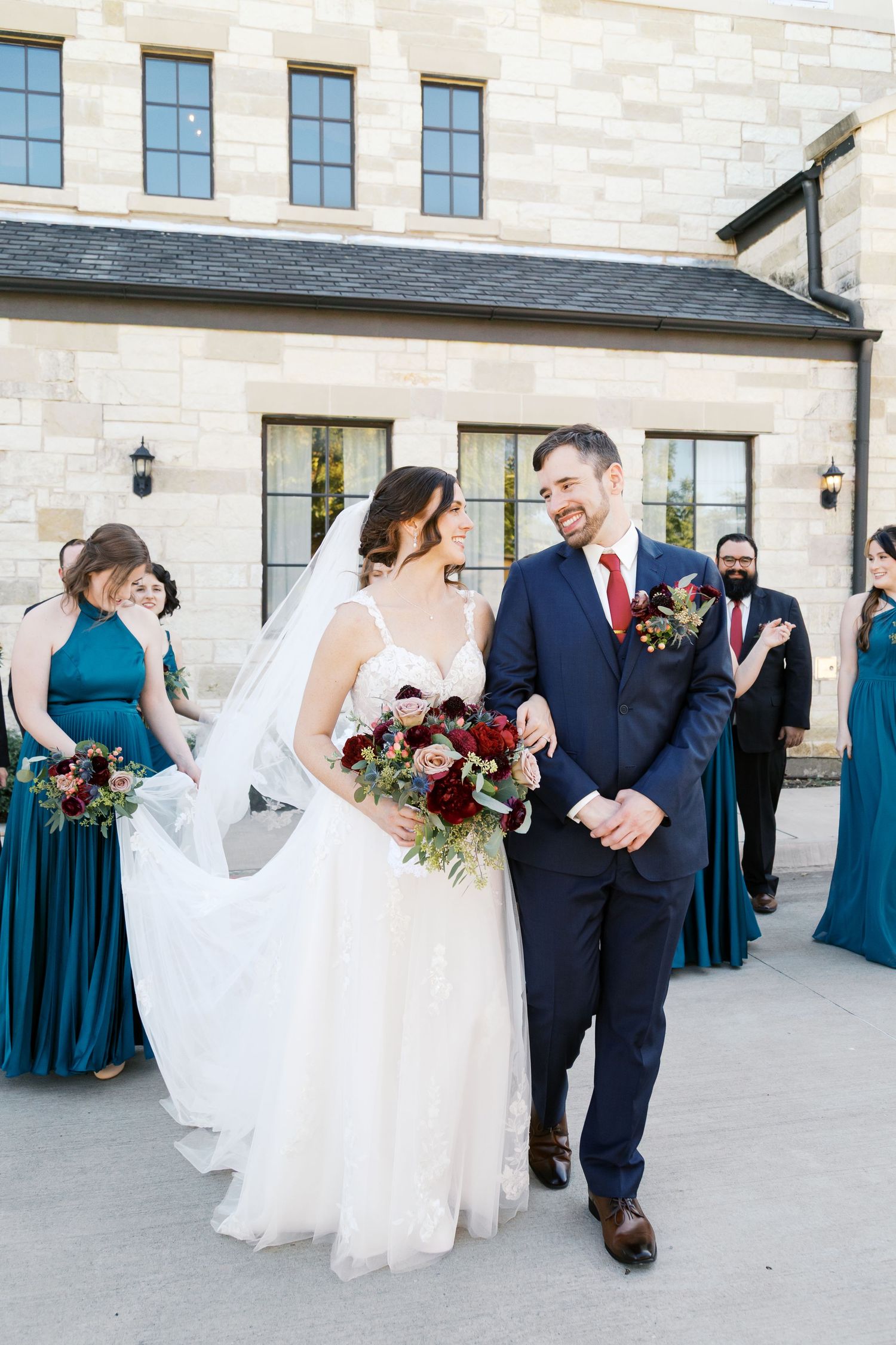 Large wedding party in teal and navy attire arranged outside stone venue.