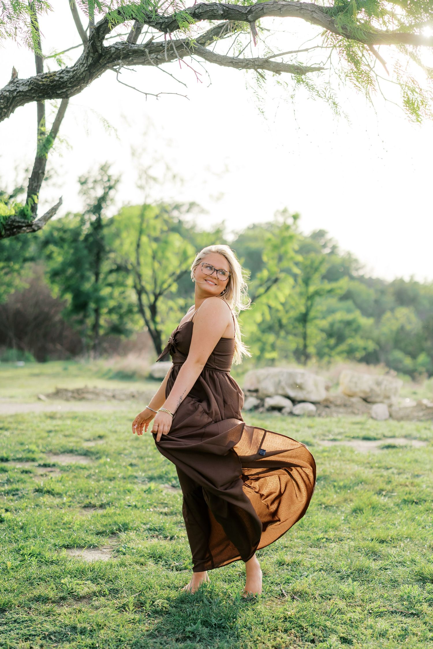 Smiling blonde woman in flowing brown dress twirling barefoot on green grass under tree in golden sunlight.