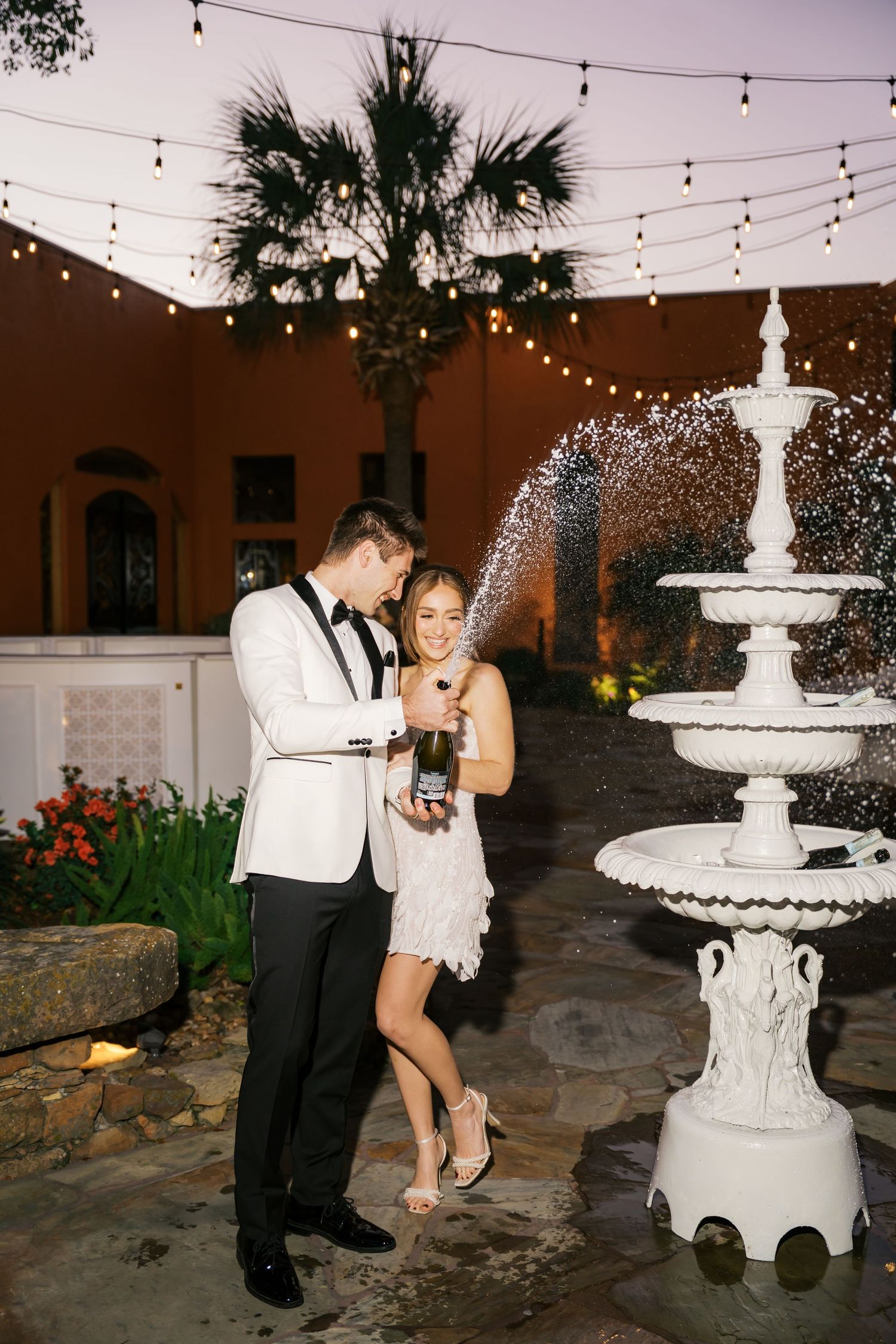 Wedding couple celebrating near an elegant white tiered fountain with string lights overhead.