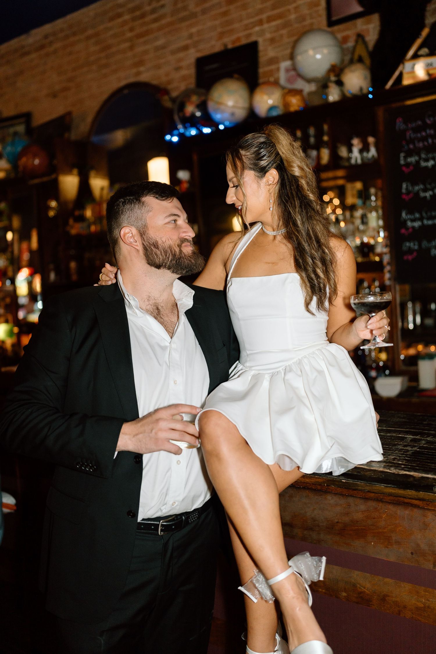Couple in formal attire shares playful moment at vintage bar counter.