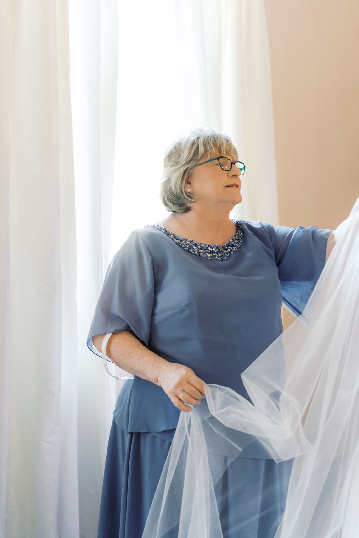 A woman in blue adjusts white curtains in a sunlit room.
