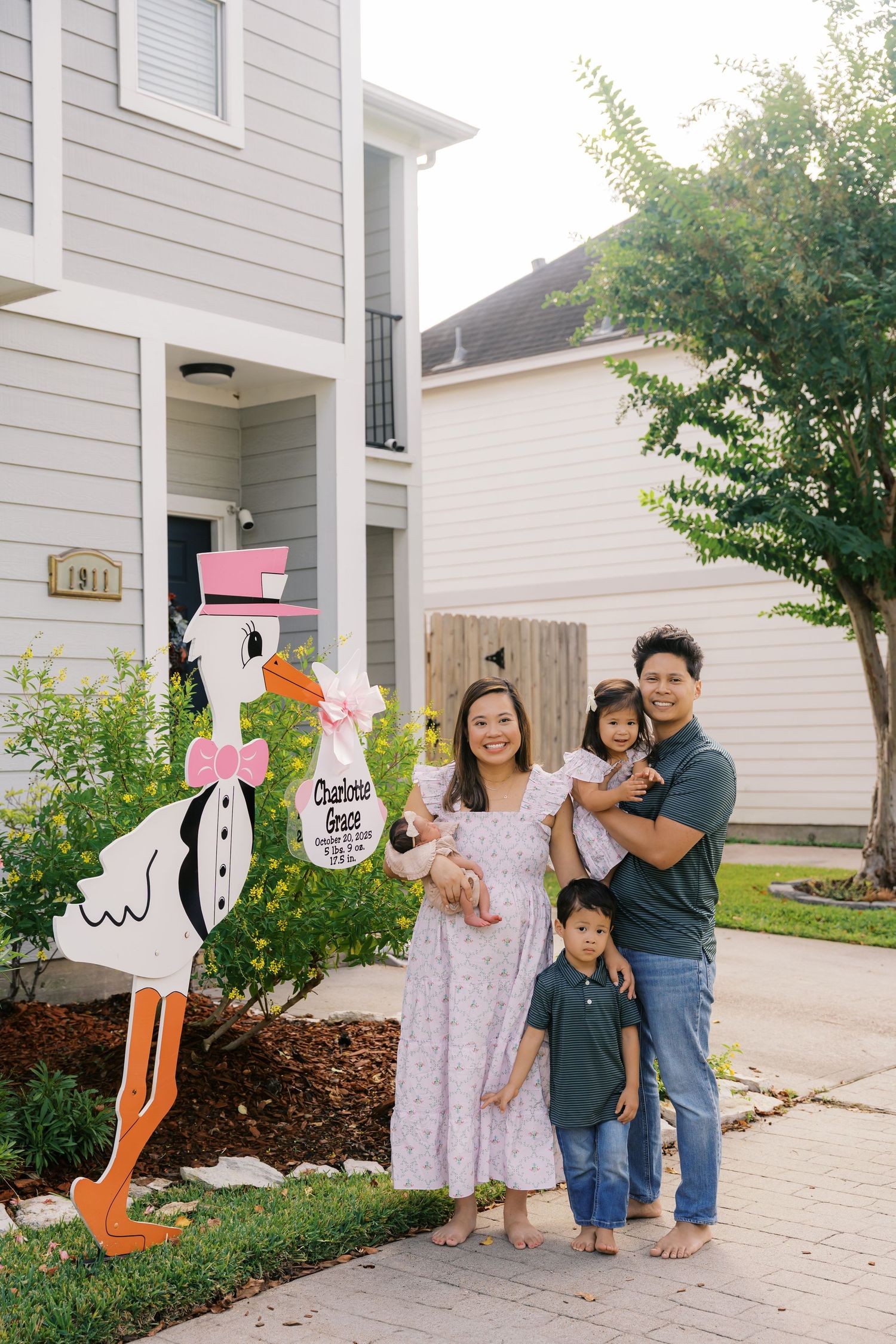 Family poses by decorative stork welcome sign outside their white house.