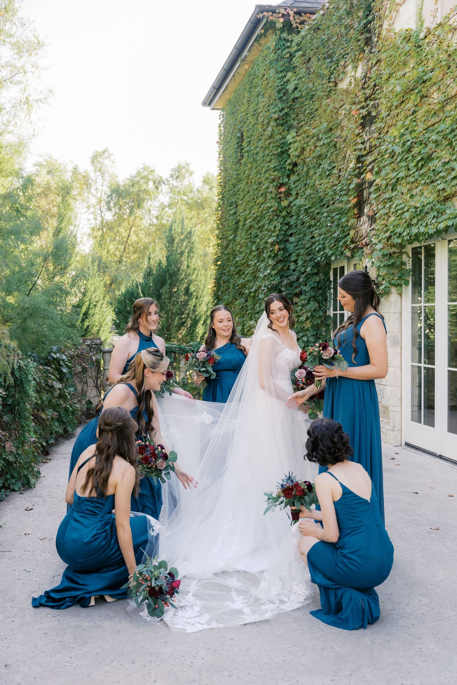 Bridesmaids in teal dresses arrange bride's long veil near ivy-covered wall.