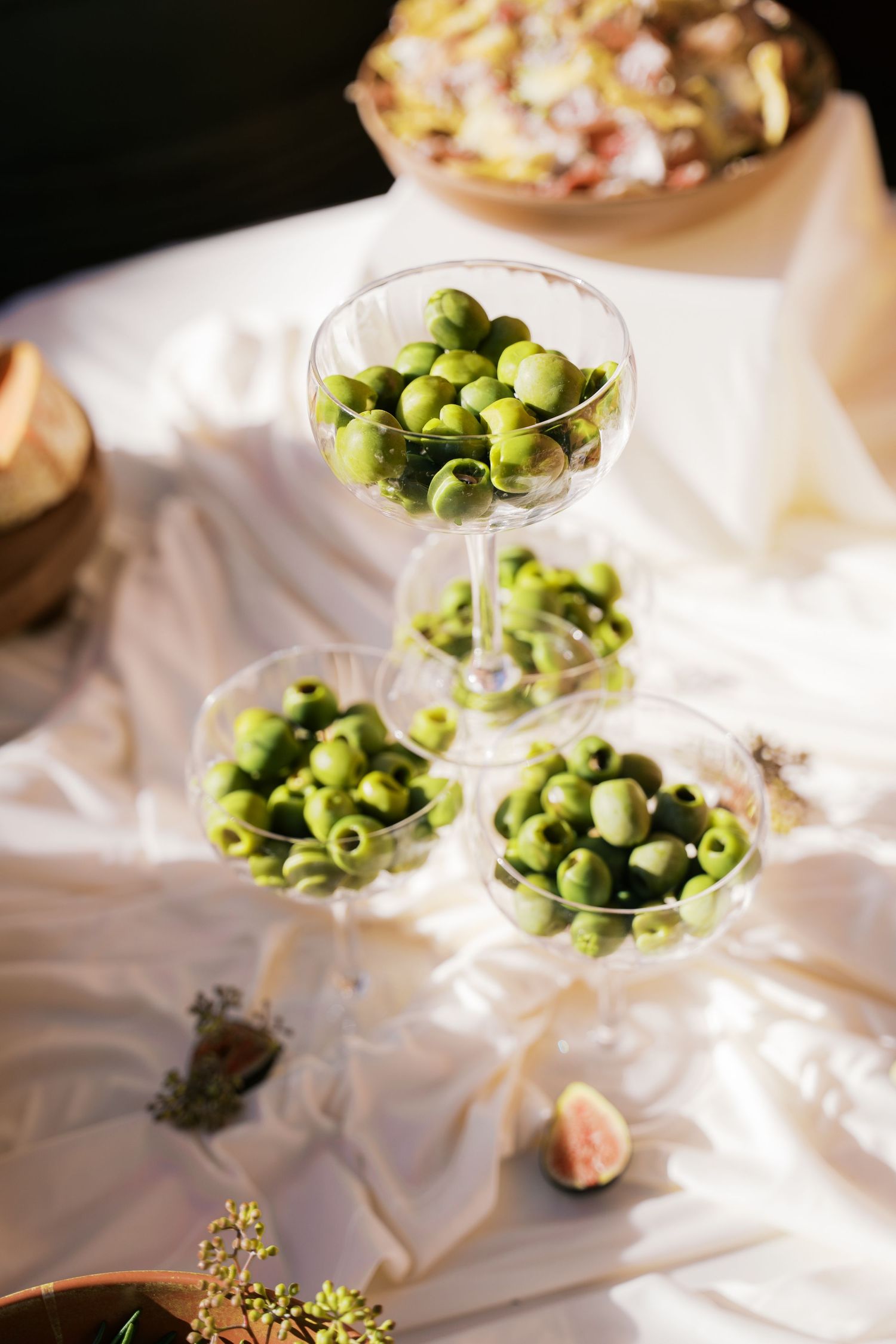 Green olives displayed in elegant glass containers on white fabric.