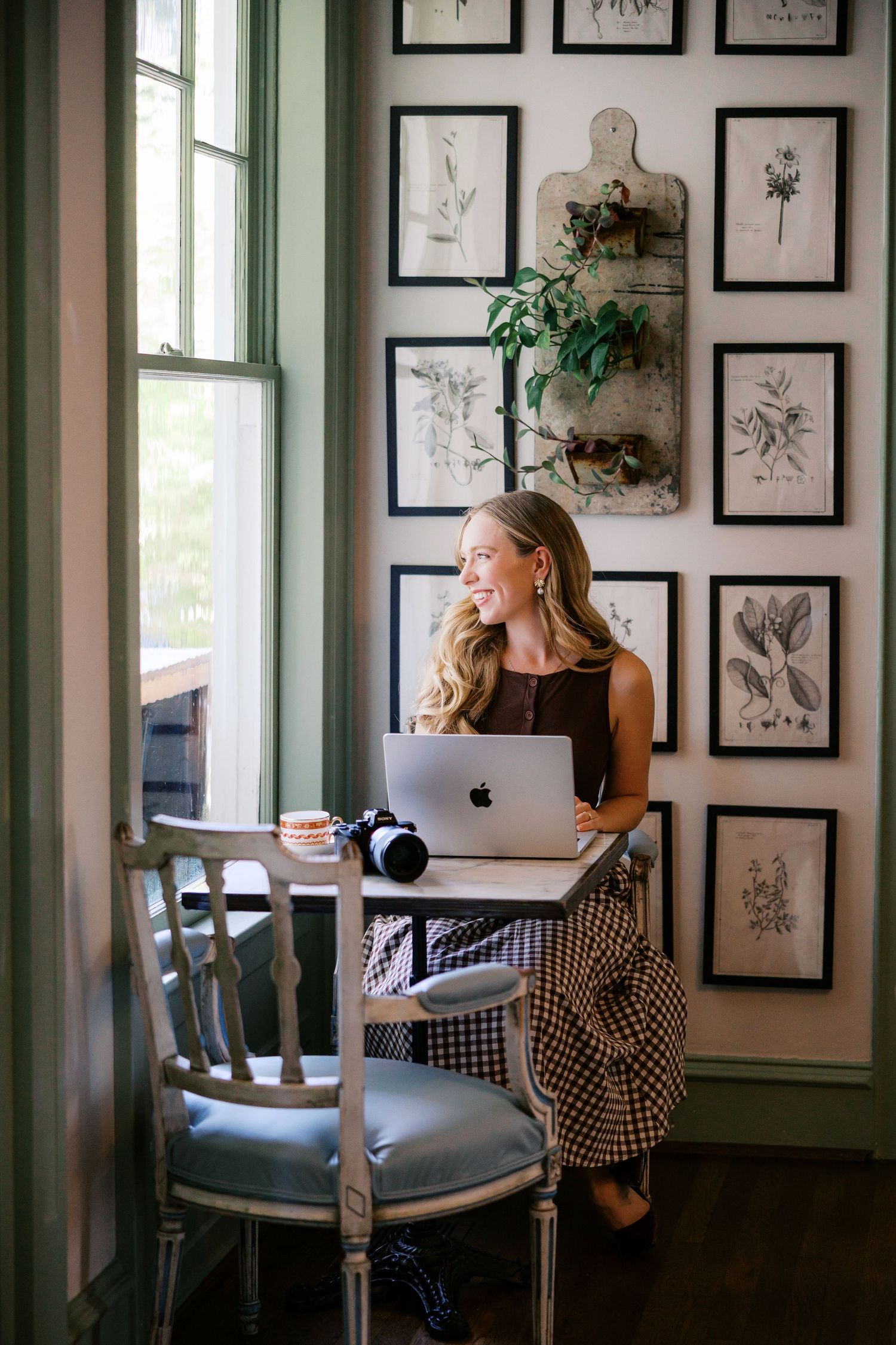 Woman smiling at window with MacBook laptop at vintage desk, botanical framed art gallery wall, camera nearby.