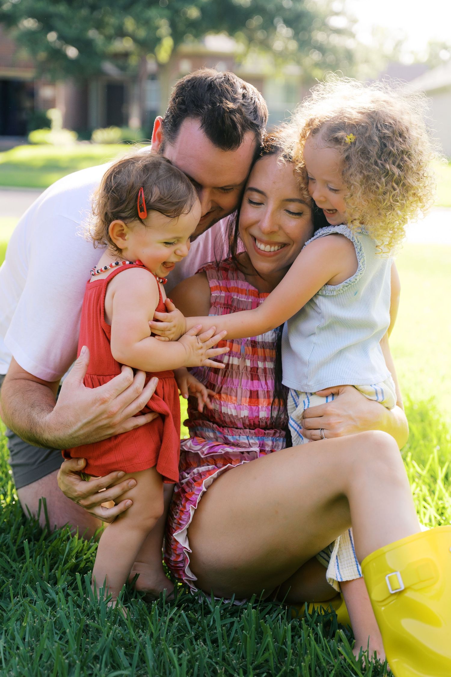 Happy family of four sitting on green grass, parents hugging two young curly-haired daughters outdoors on a sunny day.