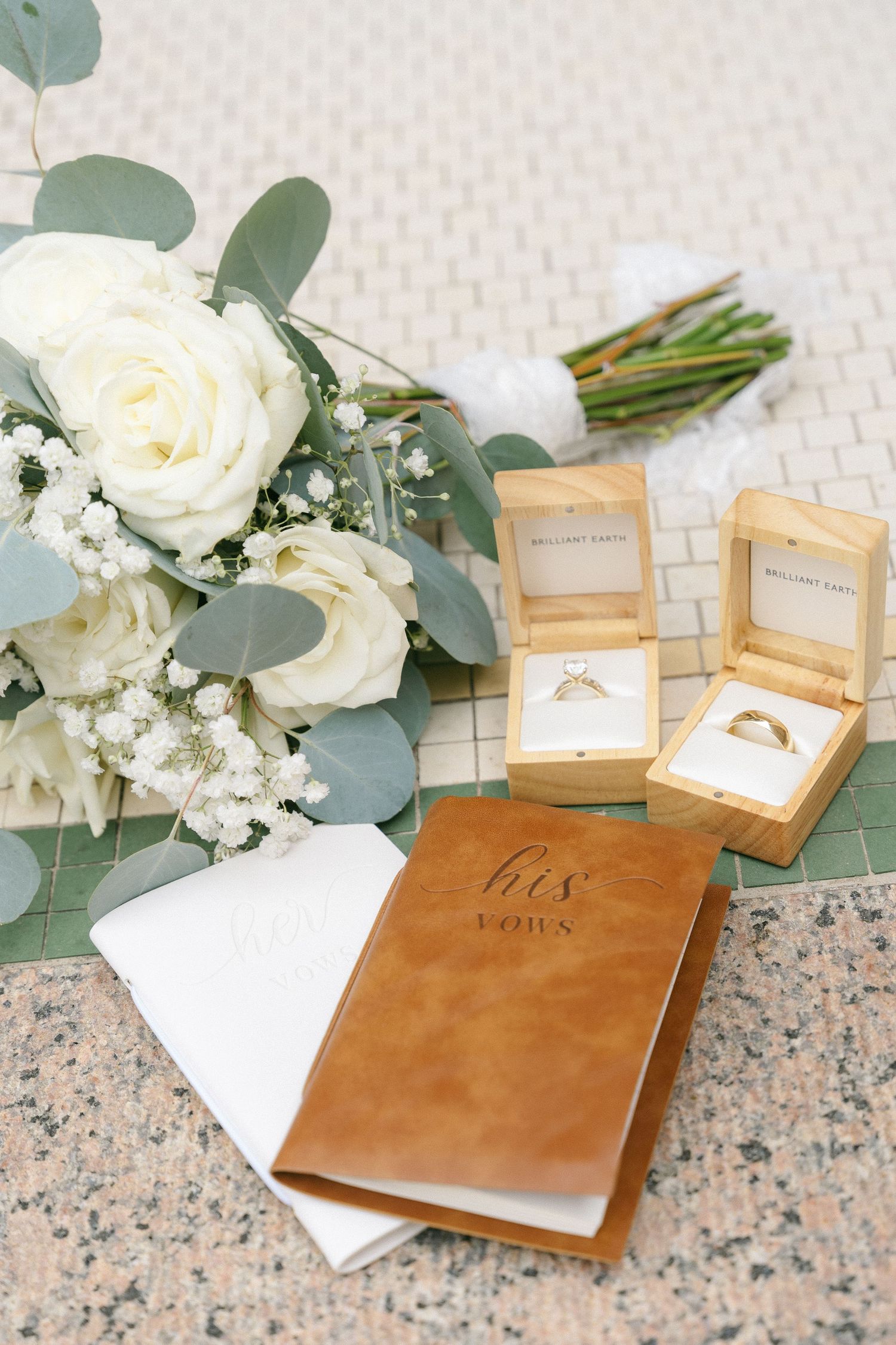 Wedding vow books and white floral arrangements with eucalyptus leaves displayed on marble surface.