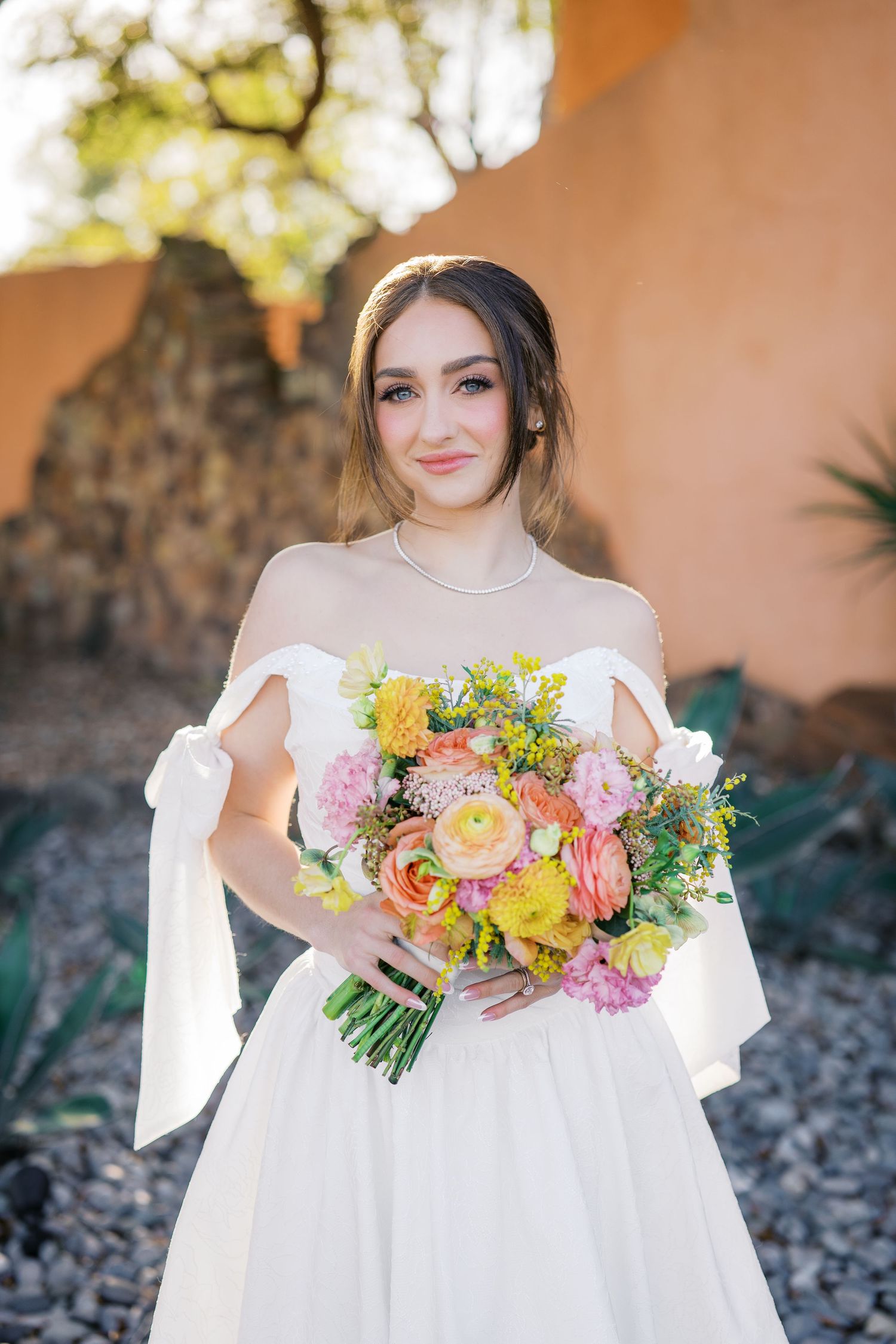 A bride in a white dress holds a vibrant bouquet of pink, yellow and orange flowers against a stone wall backdrop.