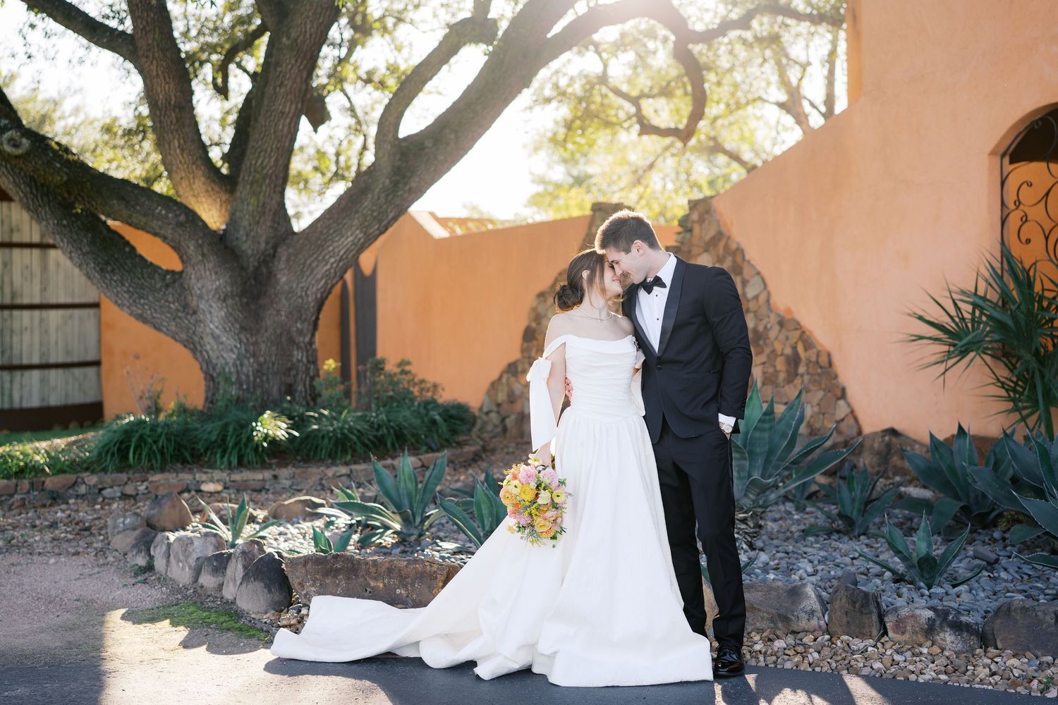 A couple poses for romantic wedding photos in front of a rustic stucco wall with trees.