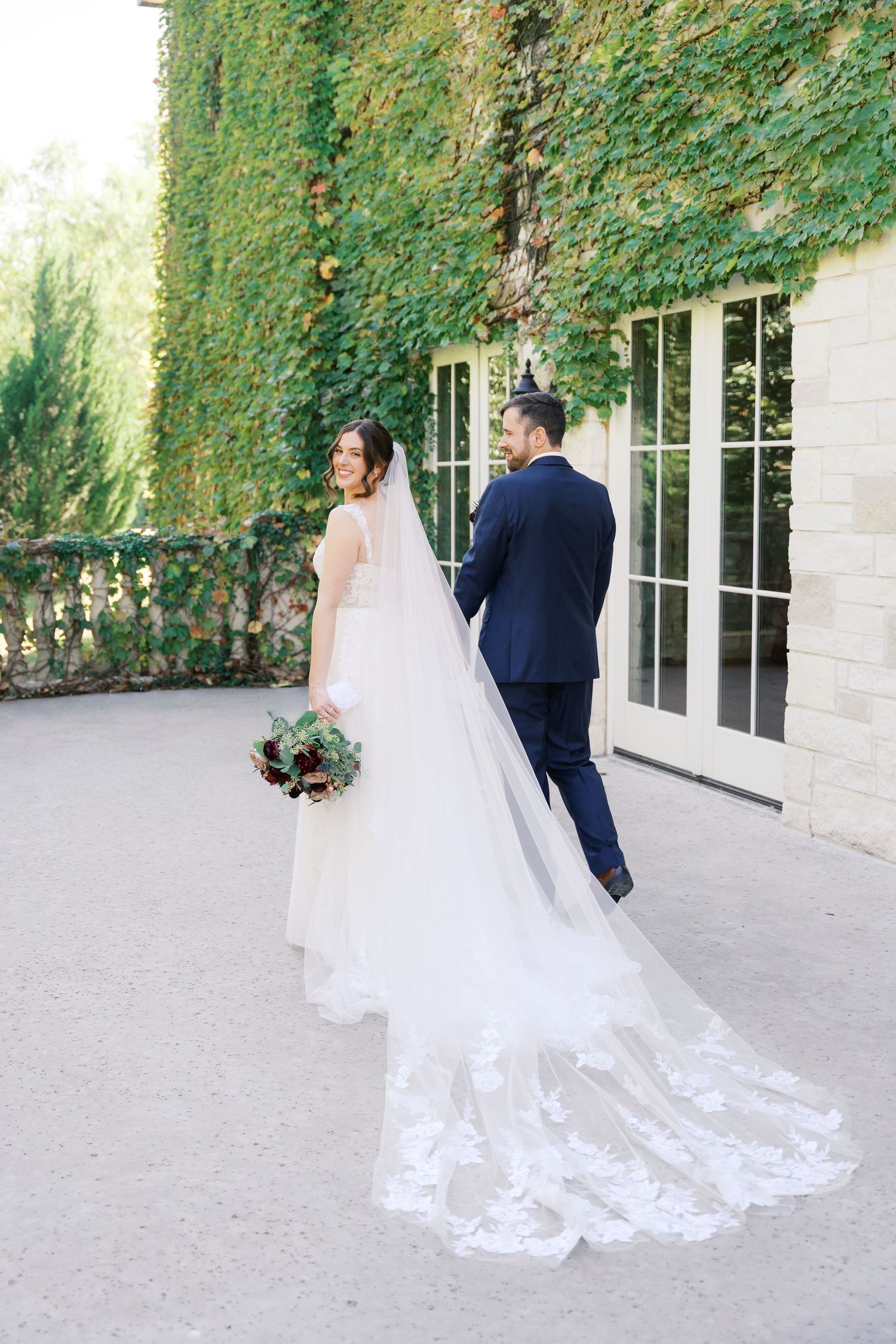 Elegant wedding couple walking along stone path with flowing white dress train and veil.
