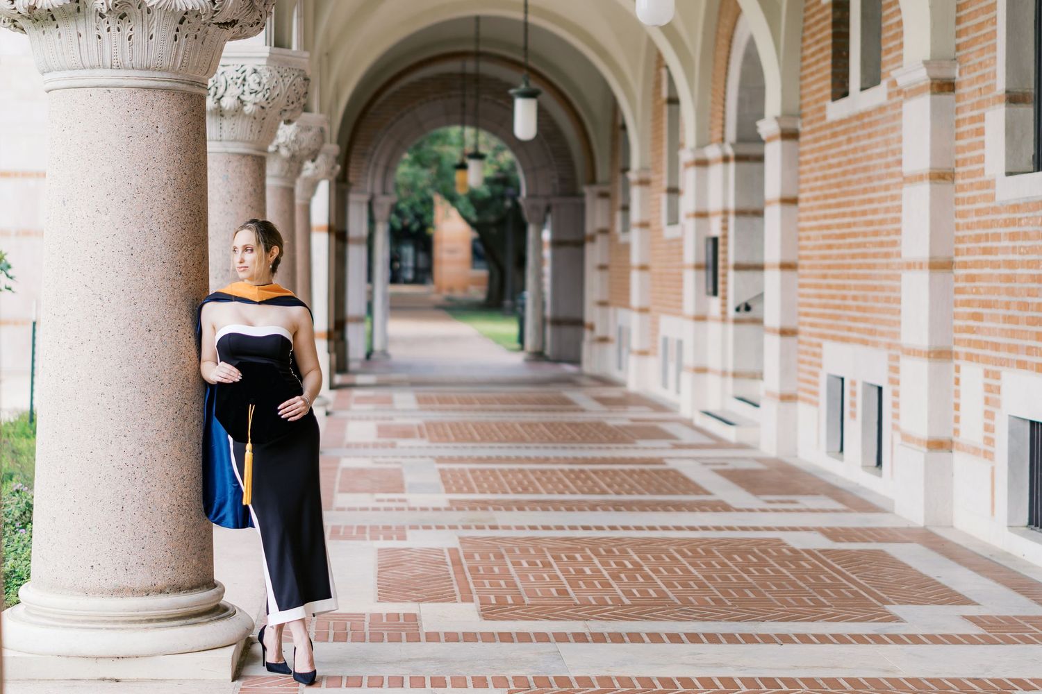Graduate in black dress poses elegantly against ornate stone column in university archway.