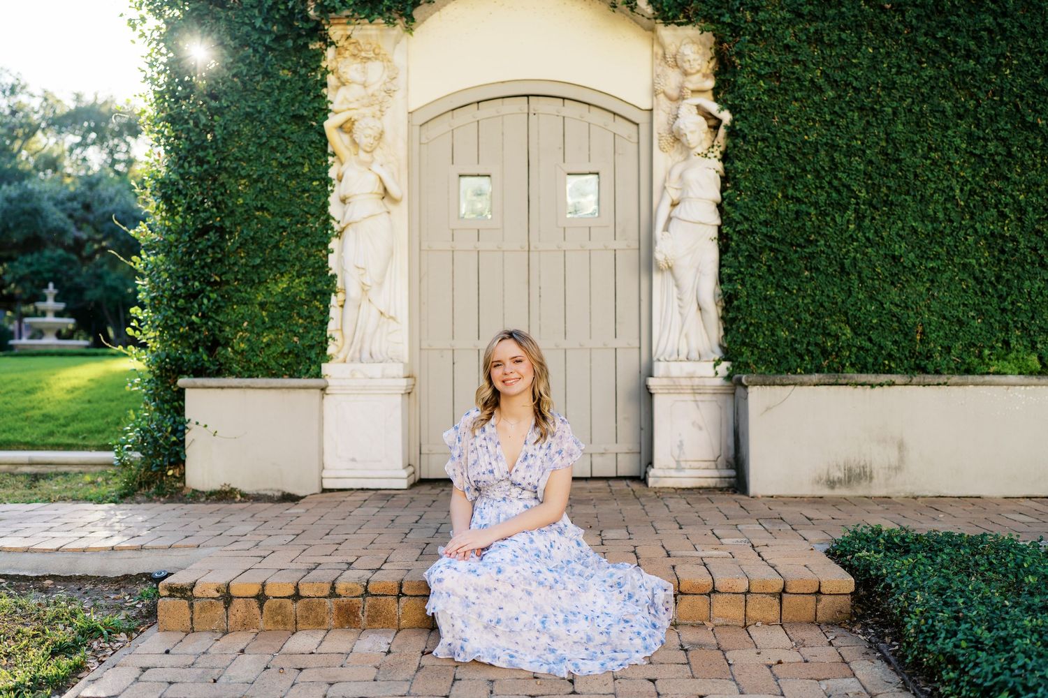 A woman in a floral dress poses in front of a stone archway and garden hedge.