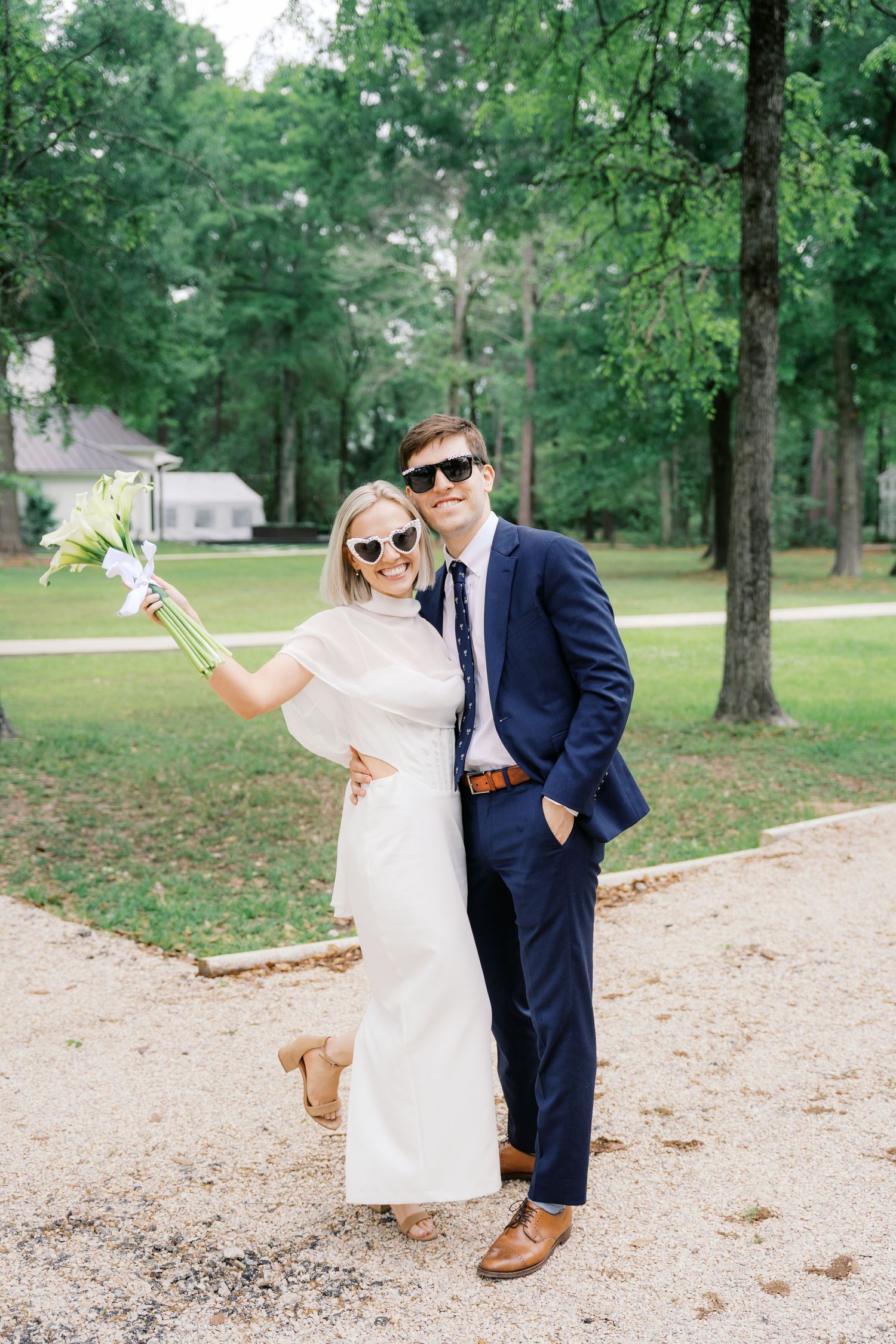 A bride in white dress and groom in navy suit wearing sunglasses pose together outdoors holding a white floral bouquet.