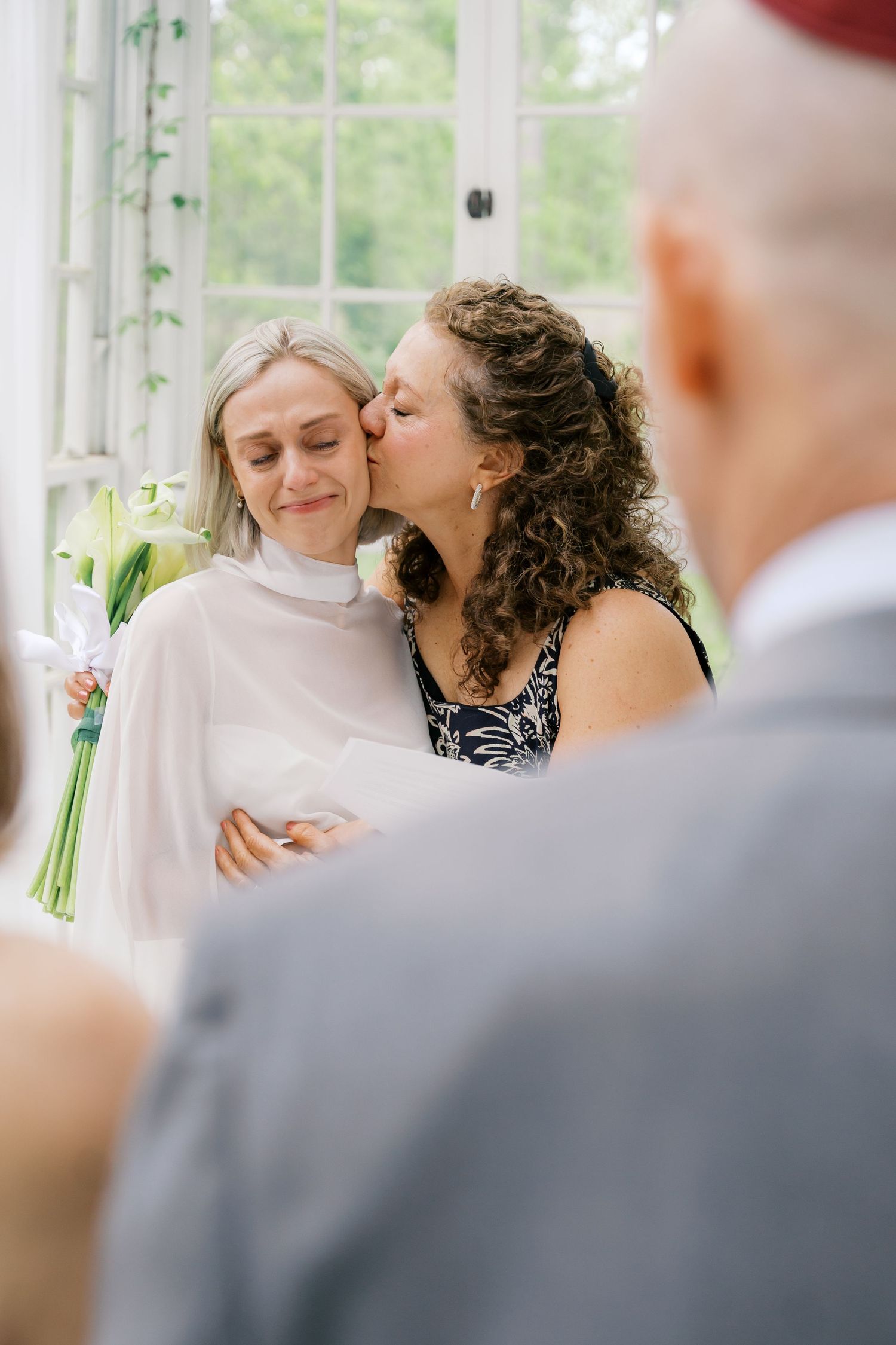 Two women share a tender kiss during a wedding ceremony, one in a white bridal cape holding flowers.