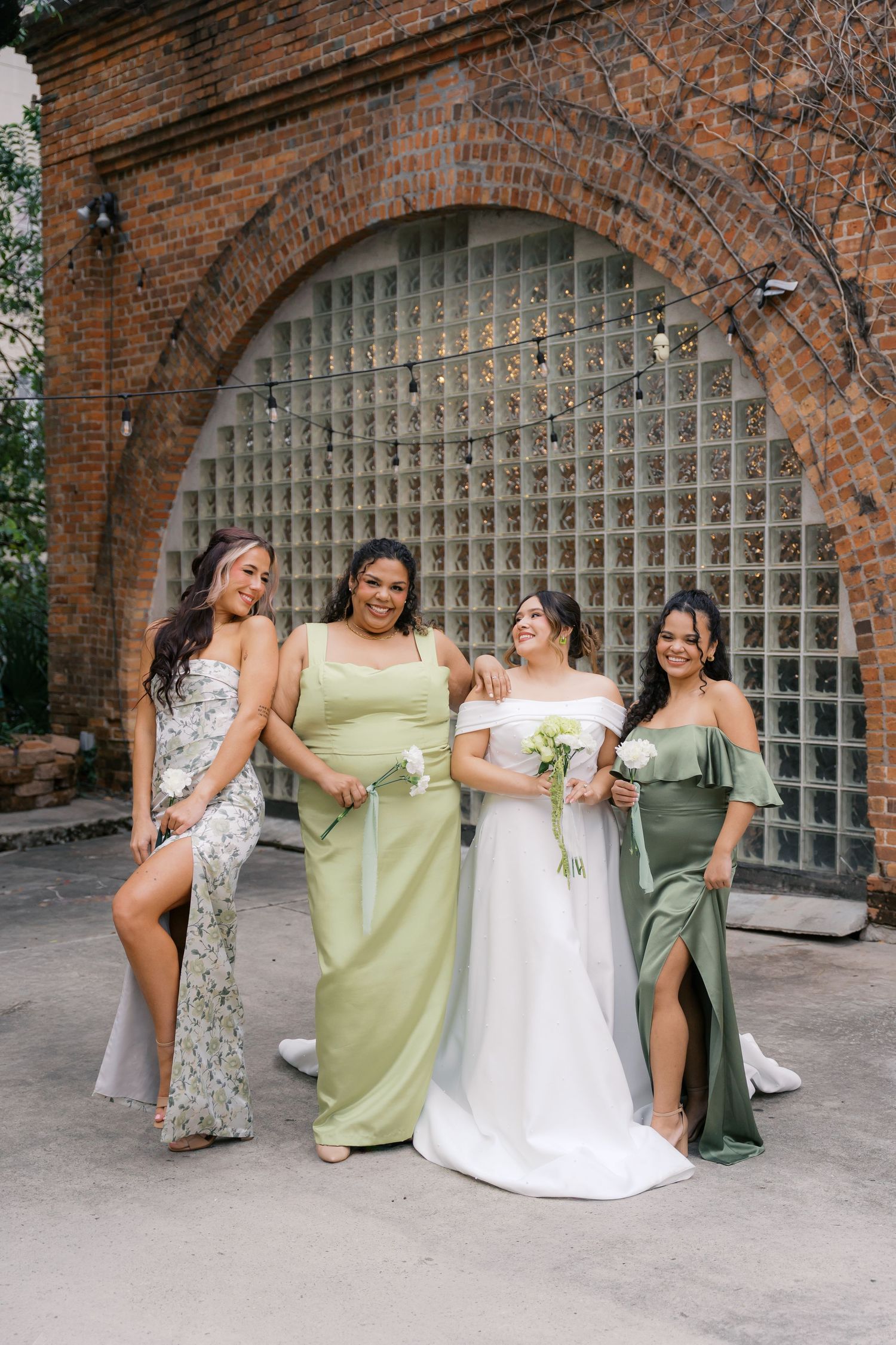 Wedding party in sage green dresses and white gown pose playfully under brick archways.