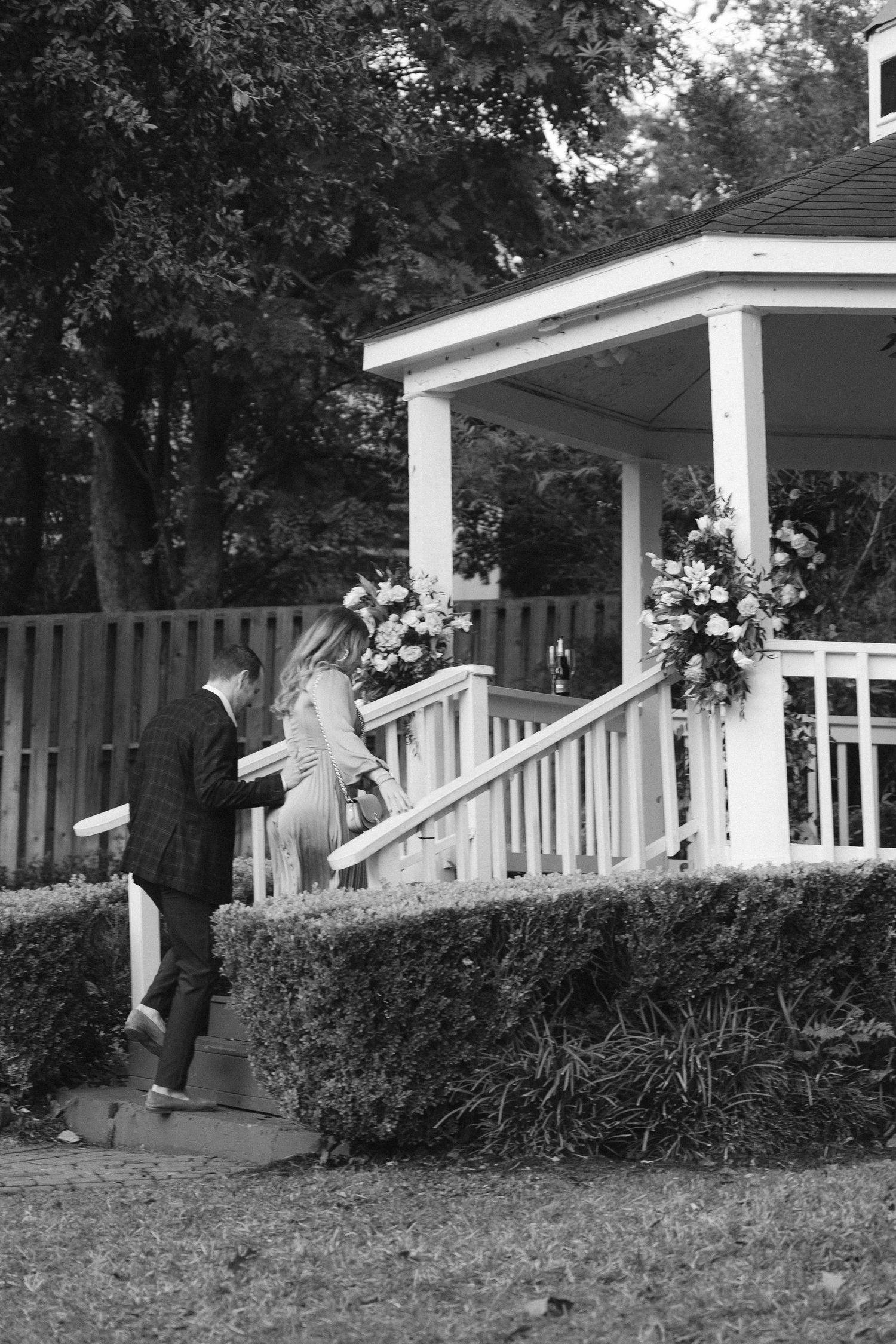 A romantic proposal moment captured in black and white on a white gazebo with wooden steps and landscaping.