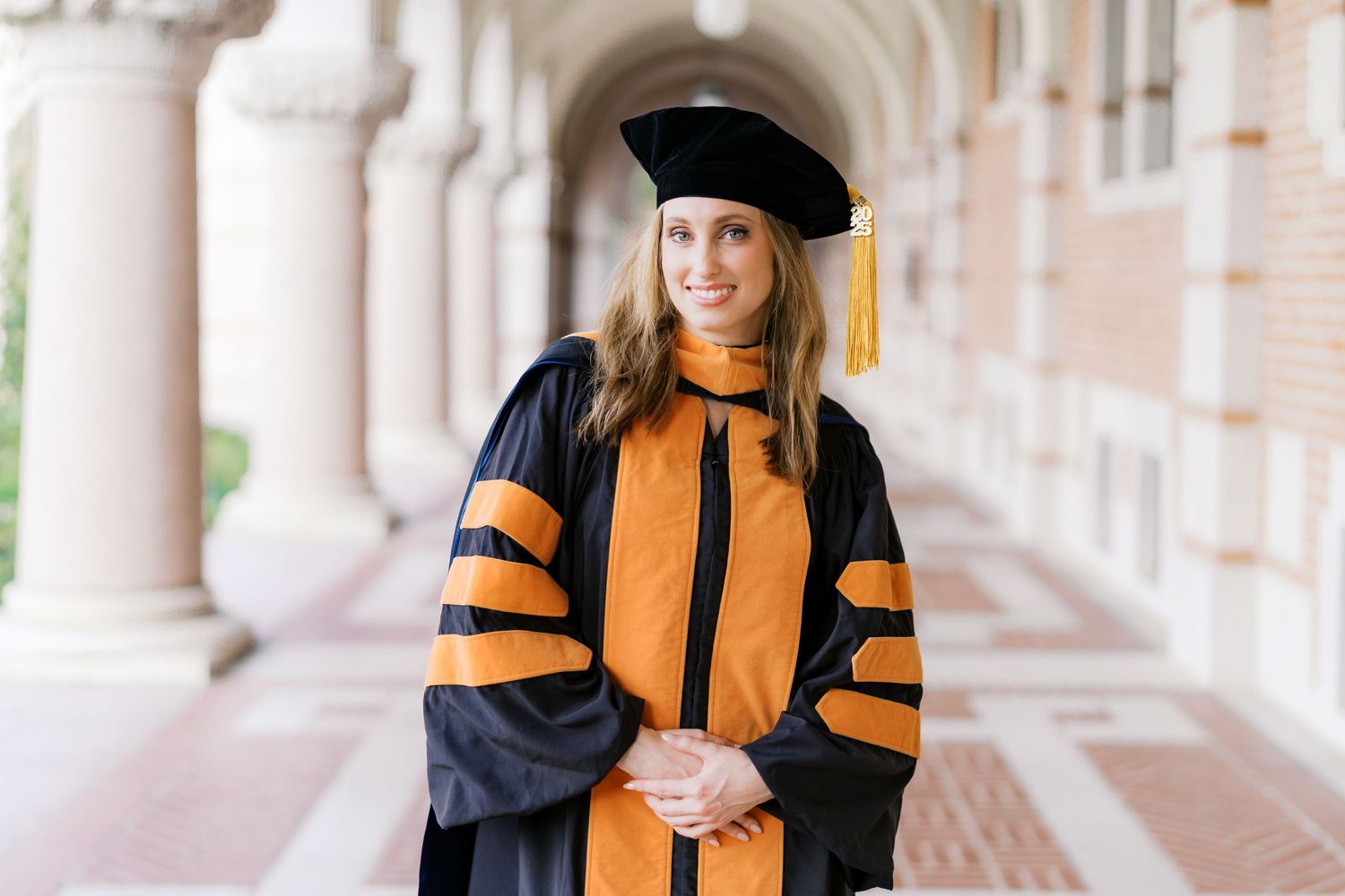 Graduate wearing doctoral regalia with orange accents poses in university colonnade.