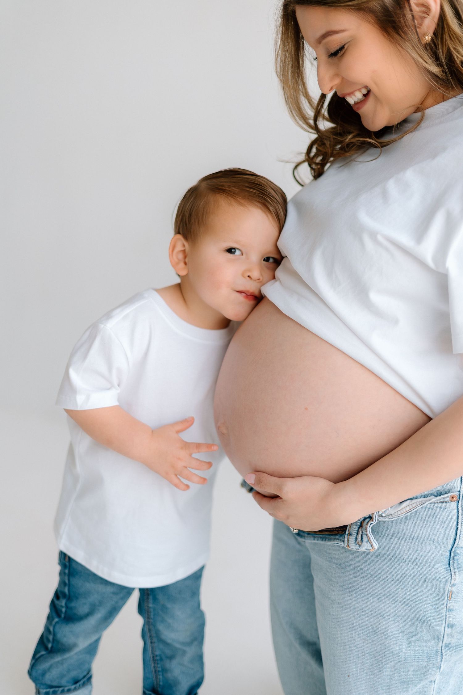A toddler in jeans and white shirt interacts with a pregnant woman's belly in a minimalist setting.