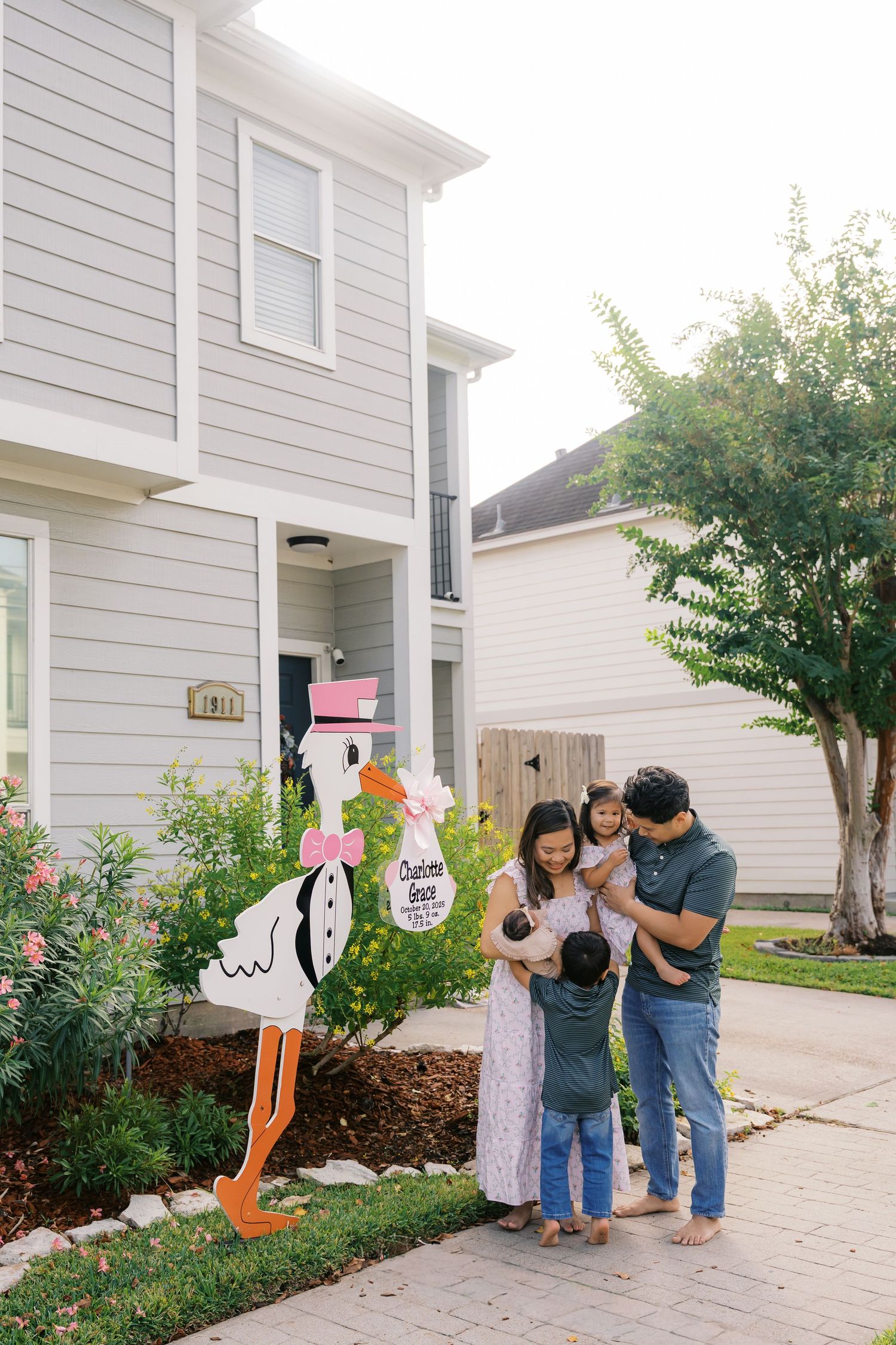 Family gathers on front porch of white house with decorative stork yard sign.
