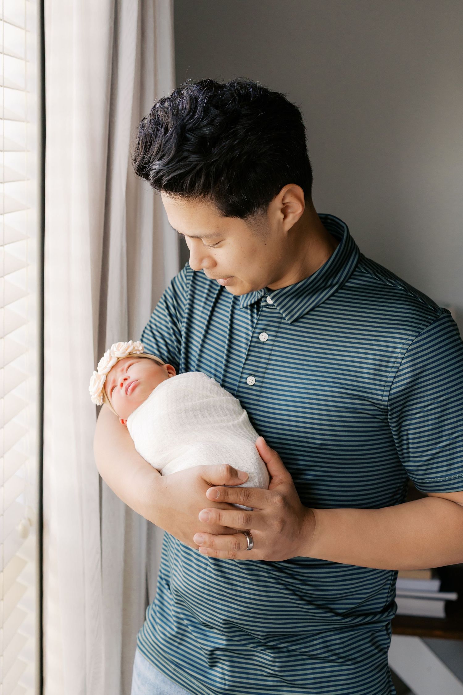 Parent in striped shirt holding swaddled newborn baby next to window with natural light.