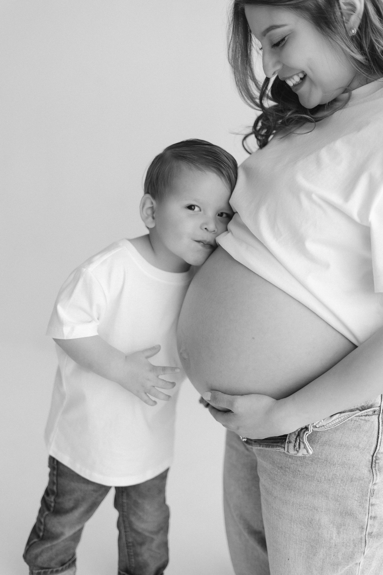 Intimate maternity photo session shows family in casual white tops and jeans against white backdrop.