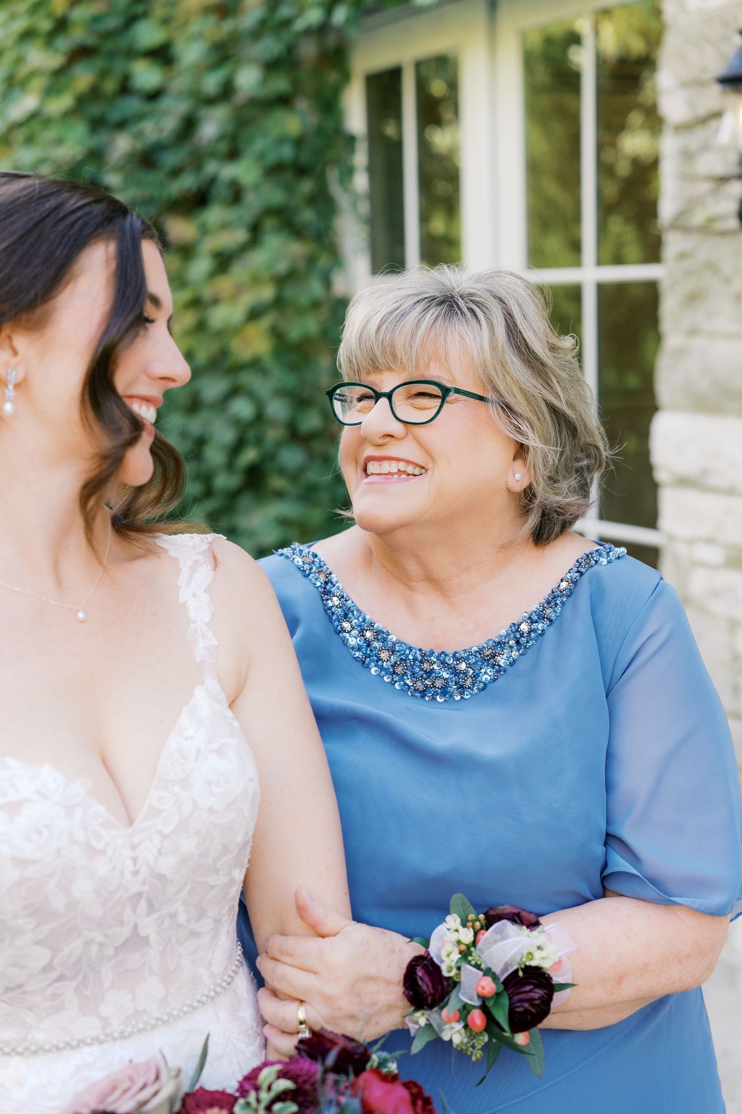 Bride in lace gown embraces guest wearing blue dress during outdoor wedding celebration.