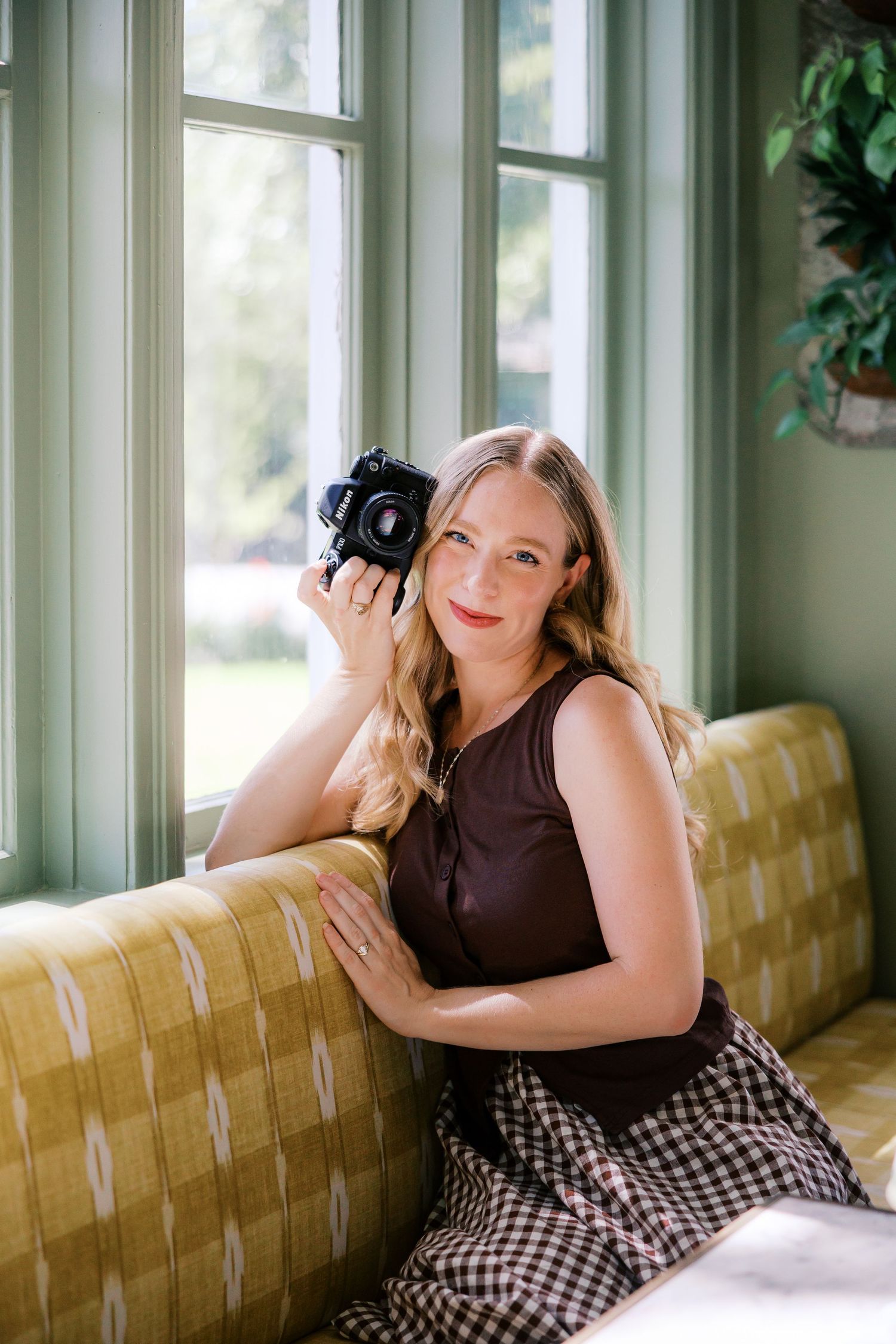 Smiling blonde woman in brown top holding a DSLR camera while sitting on a yellow sofa near green windows.
