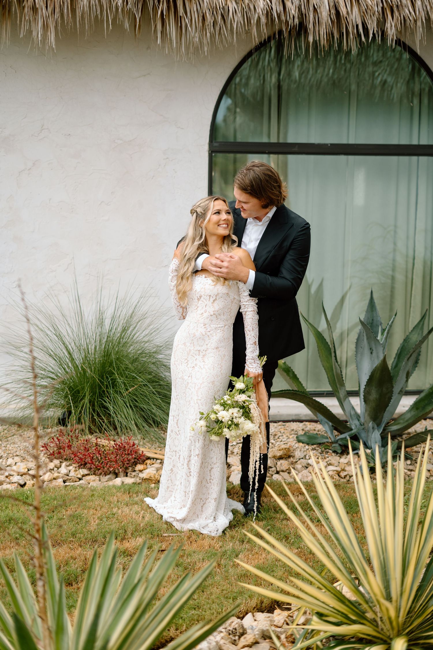 Romantic couple in formal wear embracing near desert plants and white stucco wall.