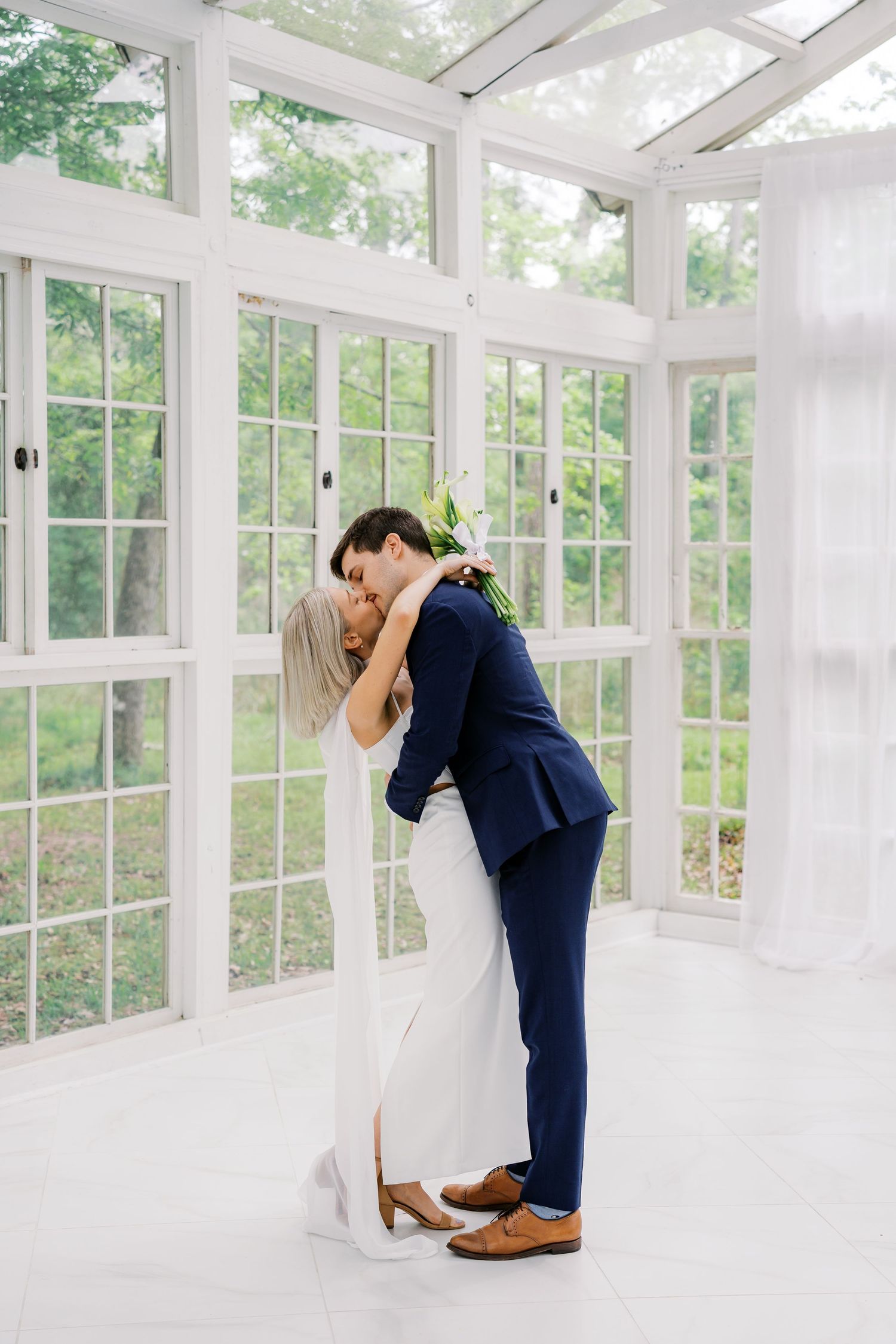 Bride and groom share a romantic kiss in a sunlit white greenhouse, bride in white gown, groom in navy suit.