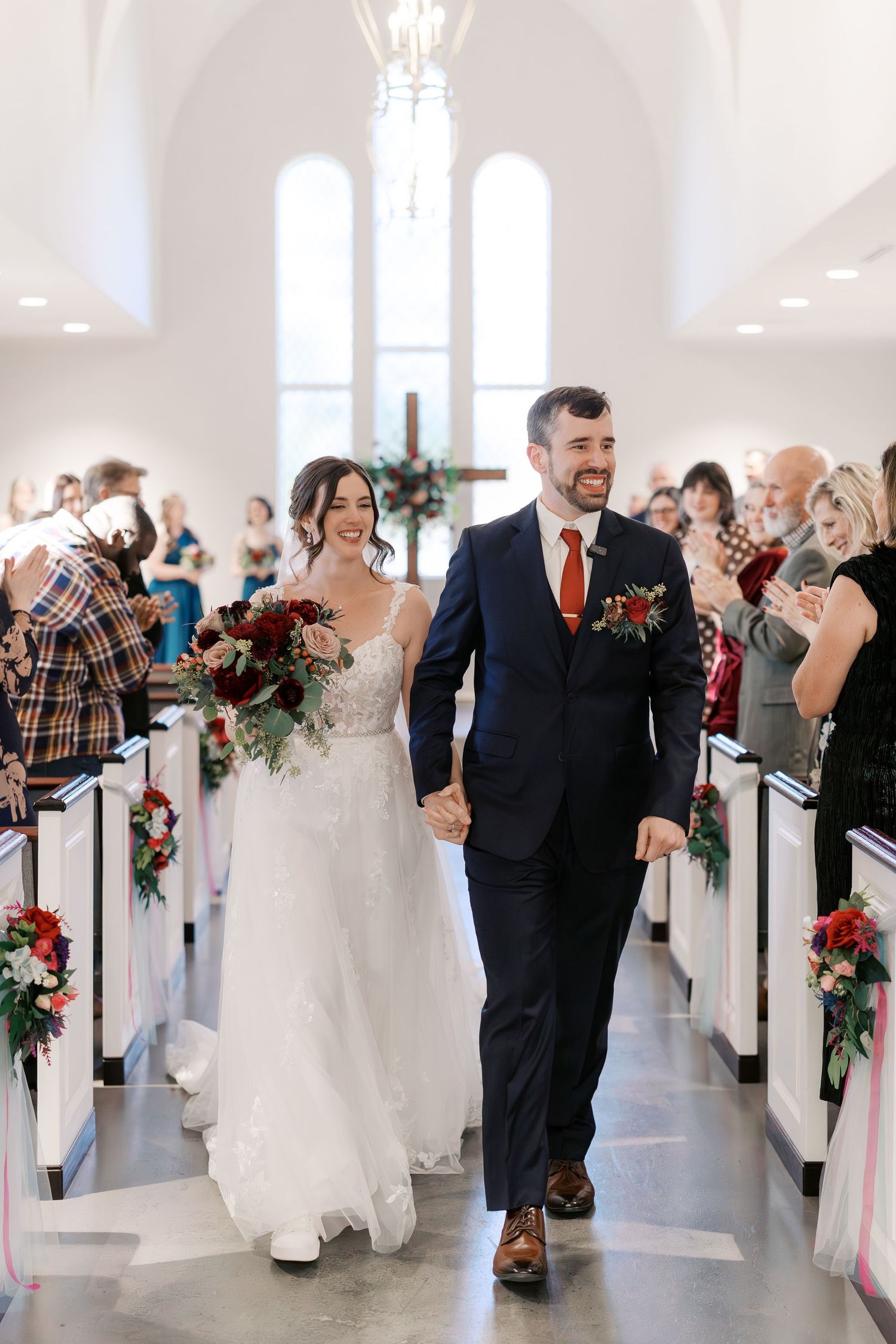 Wedding ceremony processional in white chapel with floral-decorated pews.