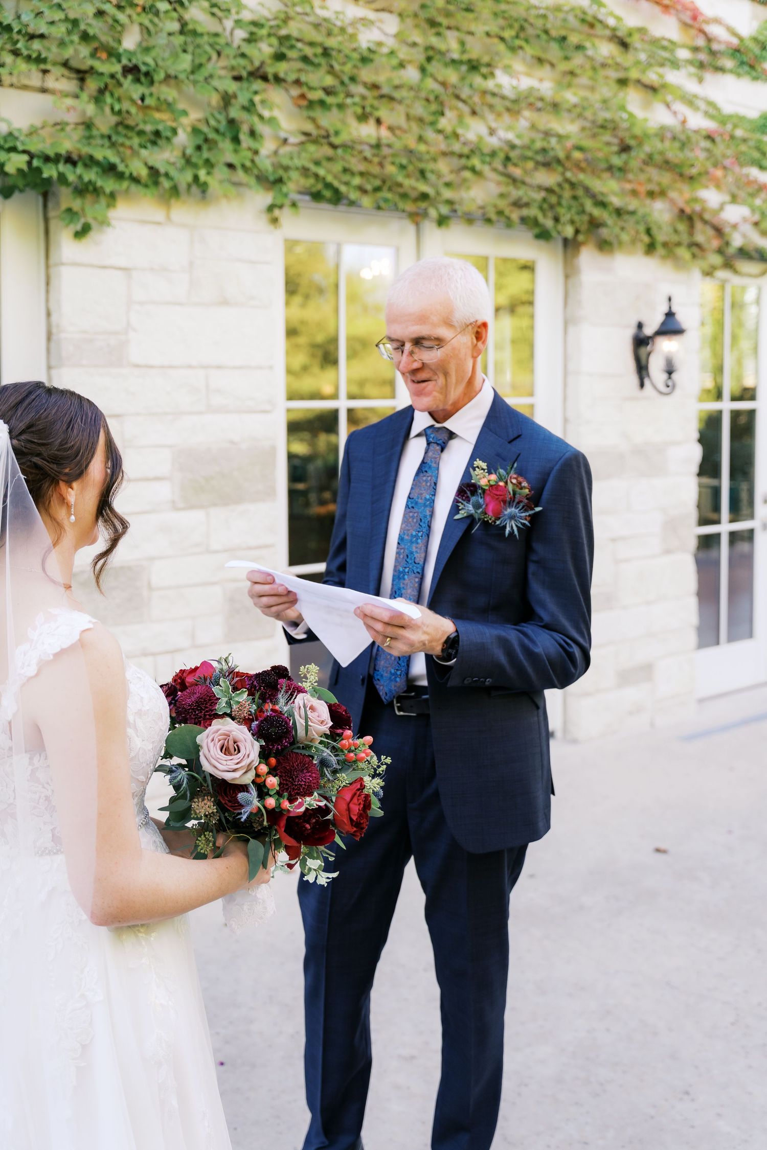 Wedding guest in navy suit reads letter while bride holds burgundy and blush floral bouquet.