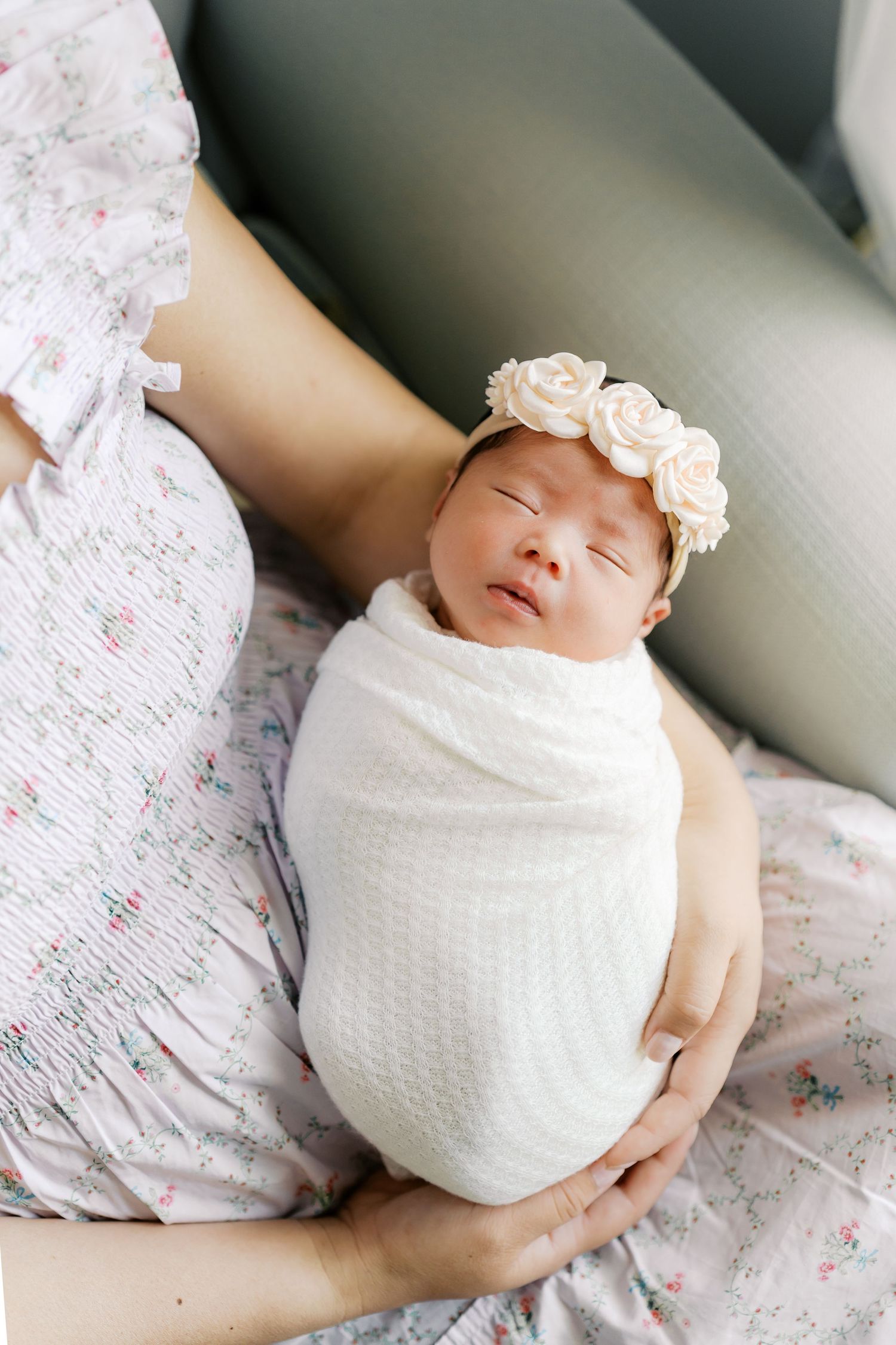 Sleeping newborn wrapped in white swaddle wearing delicate rose headband on parent's lap.
