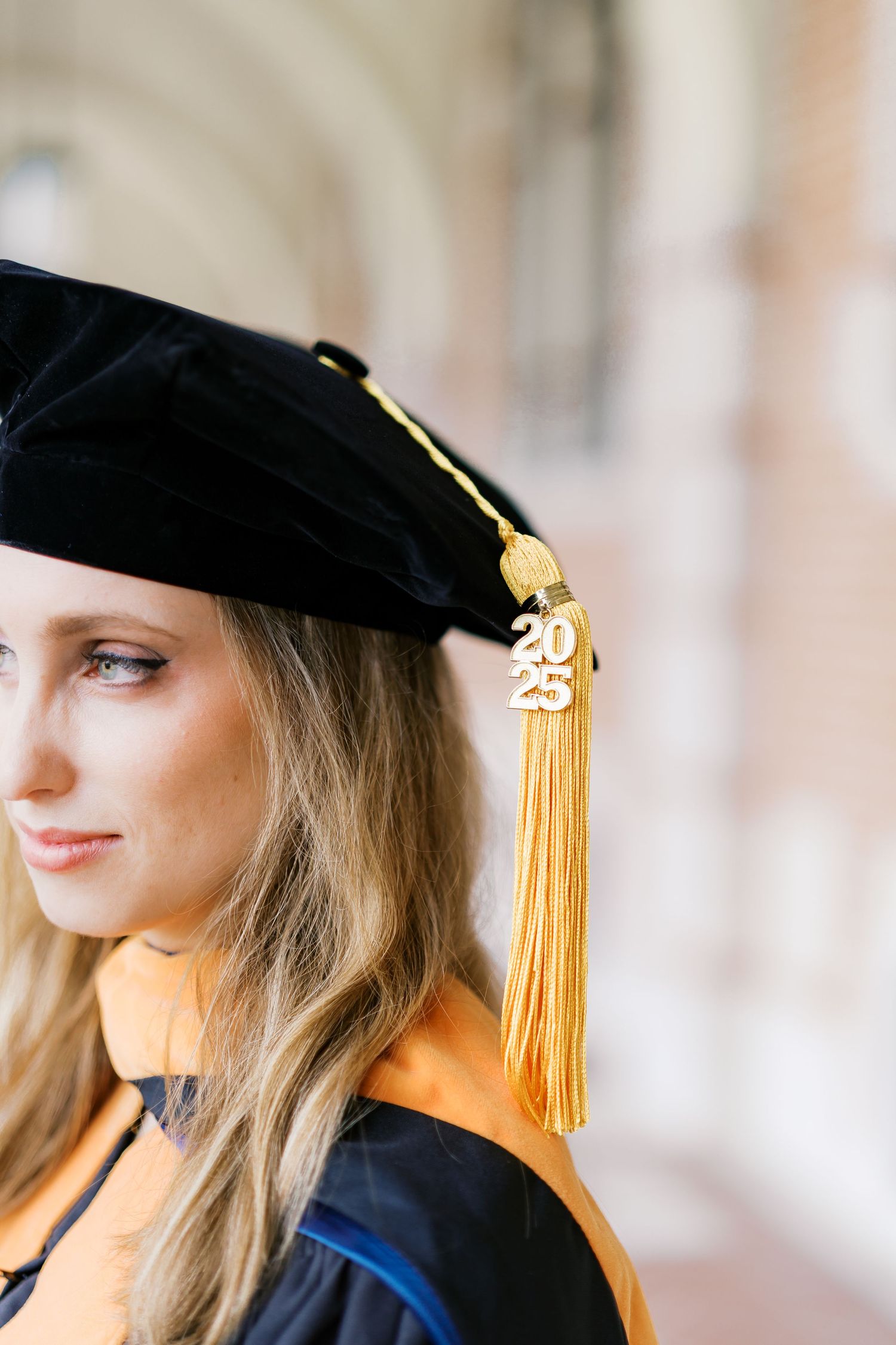 Close-up of graduation cap with gold tassel against blurred architectural background.