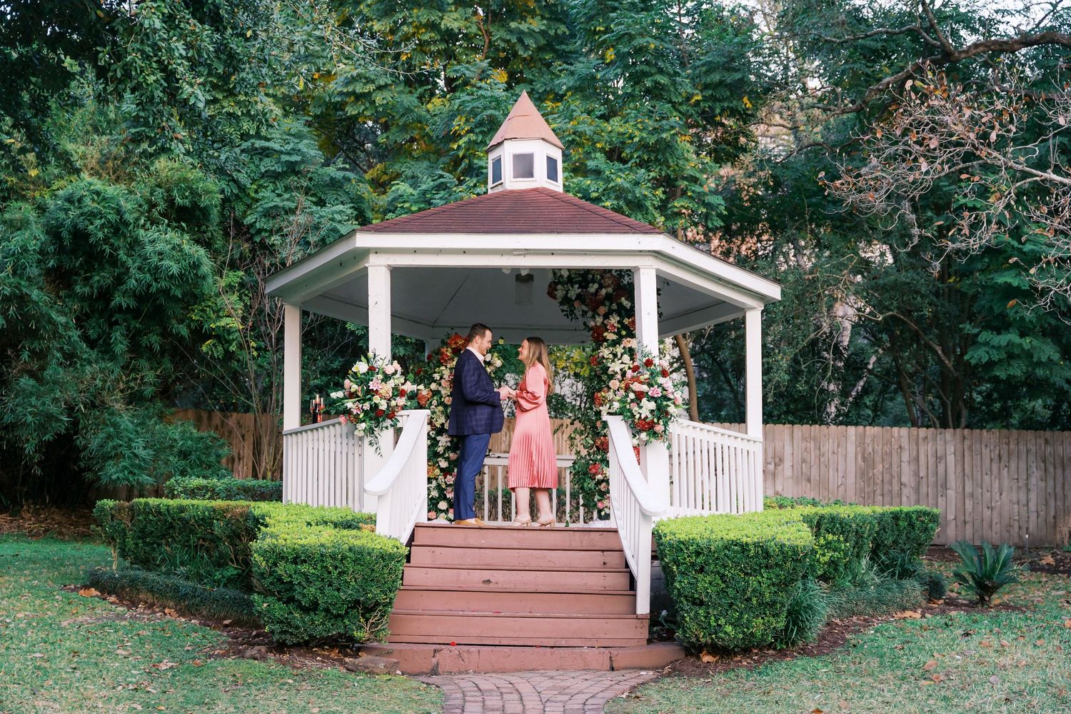 A scenic garden gazebo with brown steps and white railings surrounded by lush greenery.