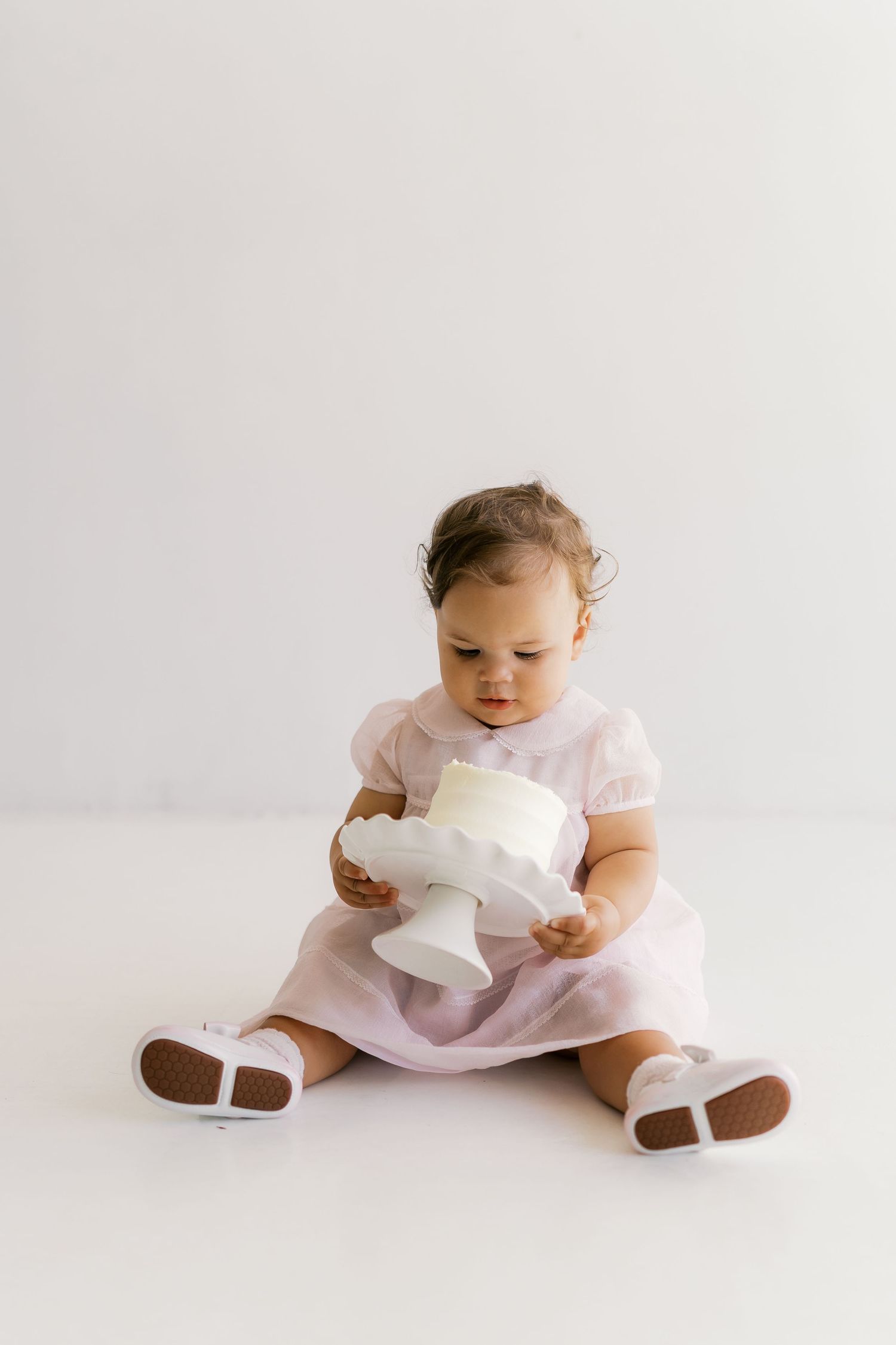 A baby in a pink dress sits on a white floor playing with toilet paper.