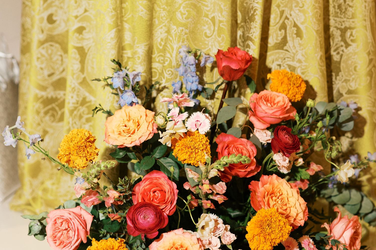 Colorful floral arrangement featuring orange marigolds, pink roses and greenery against yellow fabric.