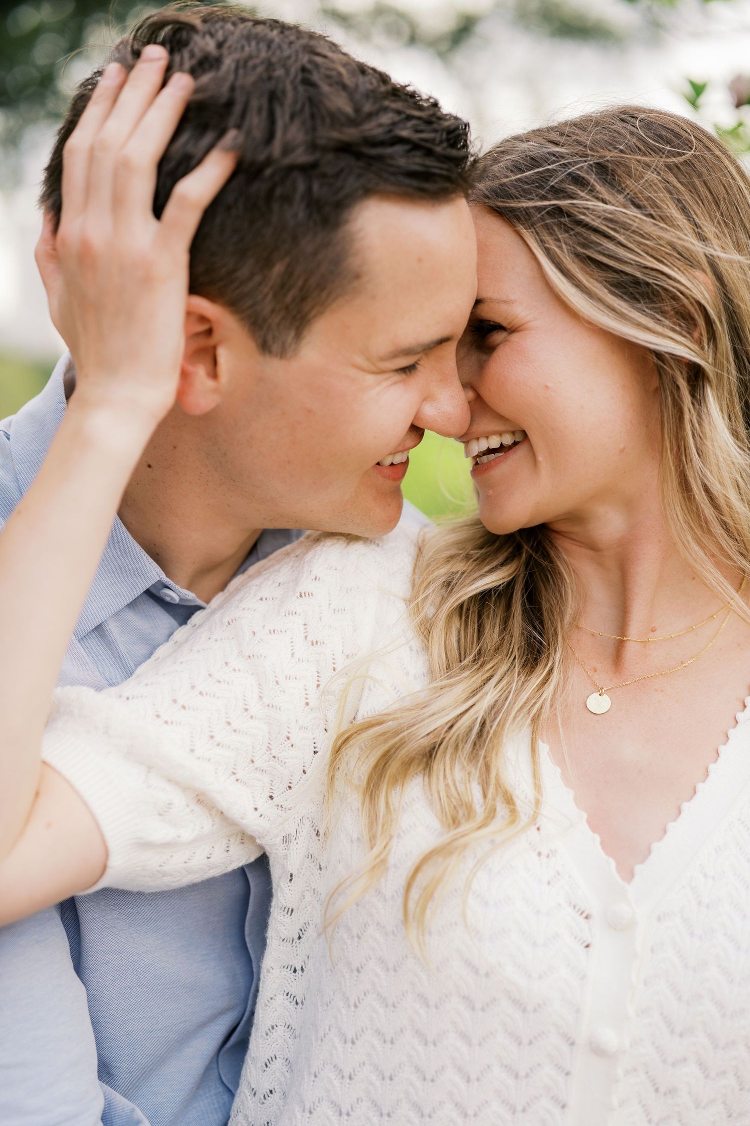 A smiling couple shares a tender forehead-to-forehead moment outdoors, woman in white knit top touching mans hair.
