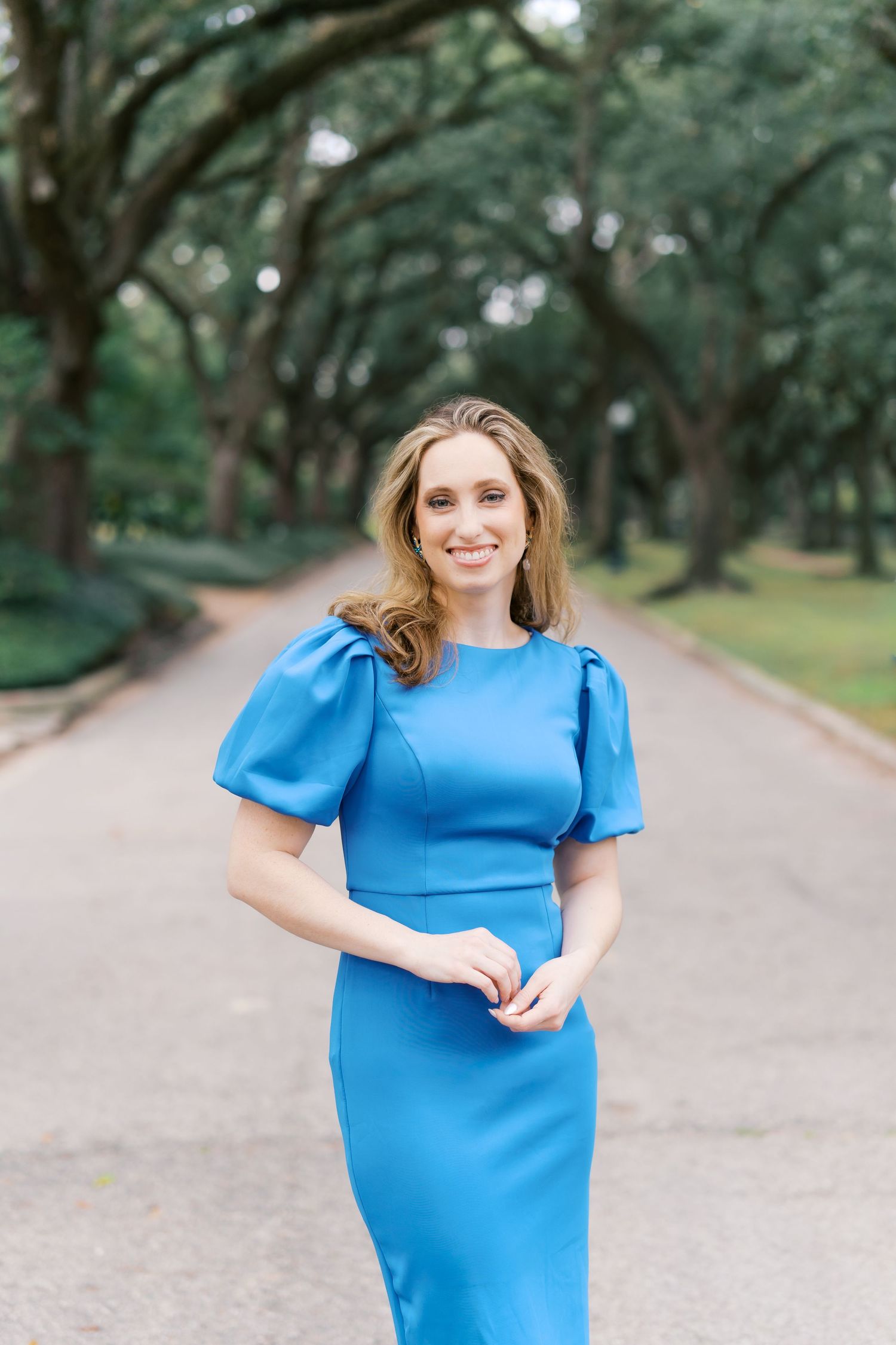 Woman in bright blue dress poses along tree-lined path during outdoor photoshoot.