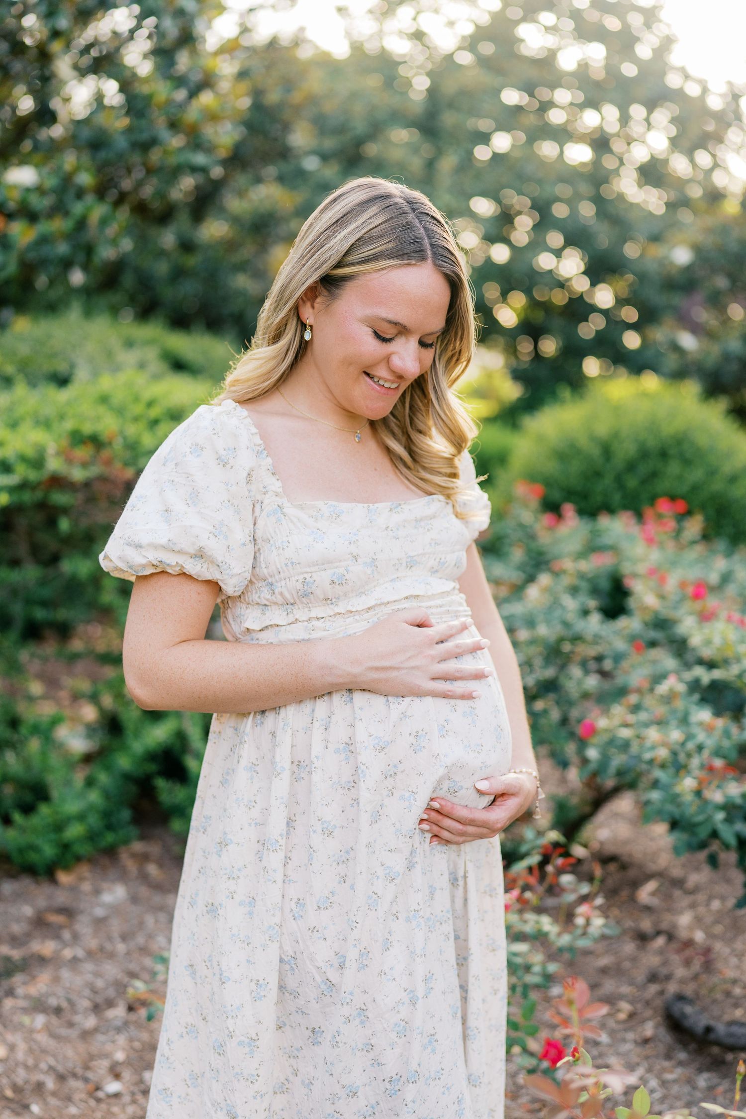 Expectant mother in white dress smiles while cradling baby bump in garden setting.