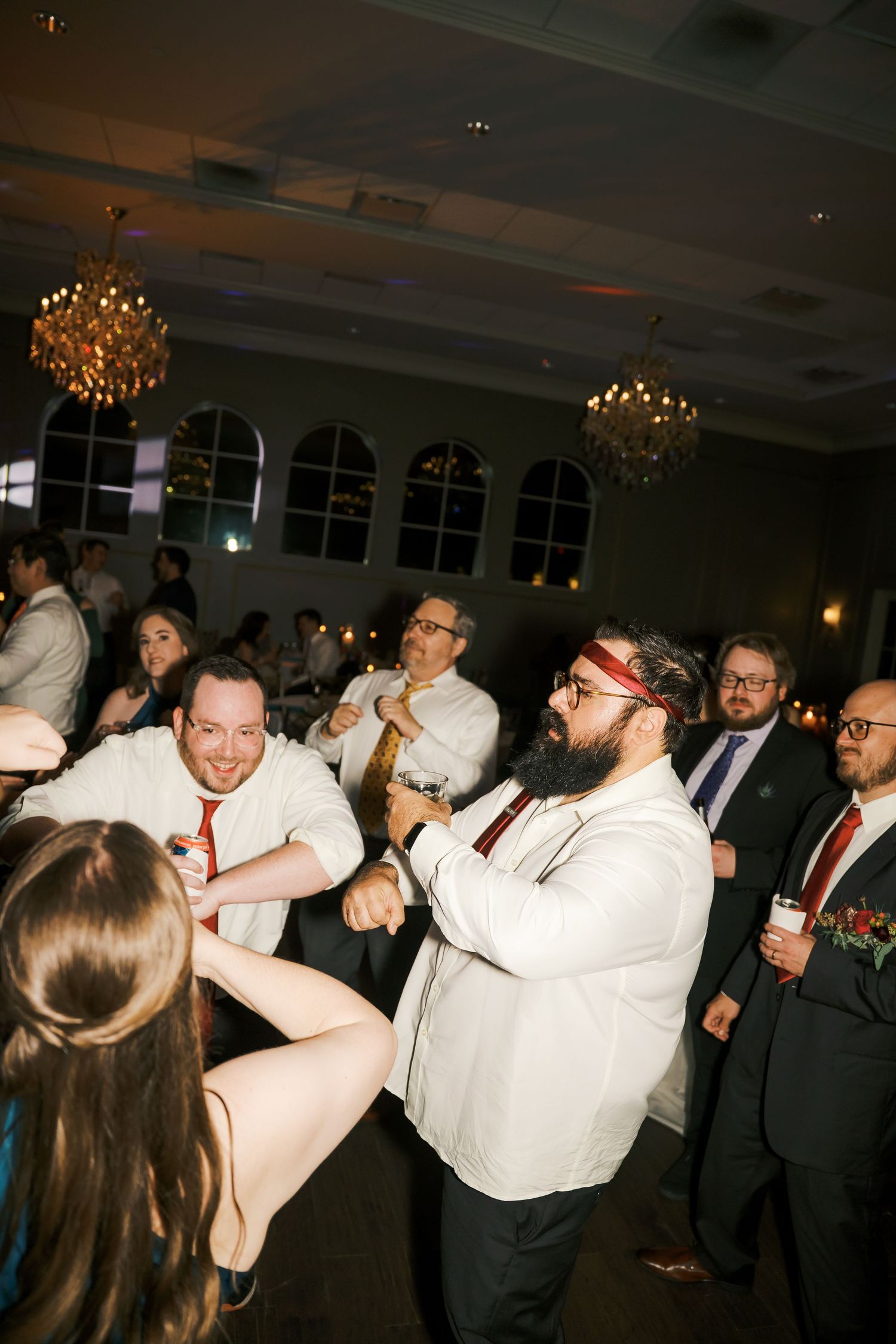 Wedding guests dance energetically on the dance floor under elegant chandeliers at an indoor reception venue.