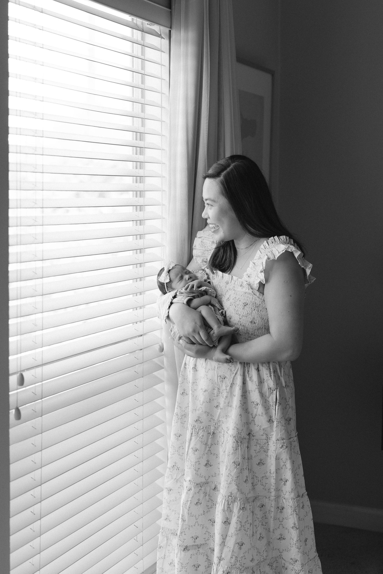 Black and white photo of mother holding newborn baby by window with venetian blinds.