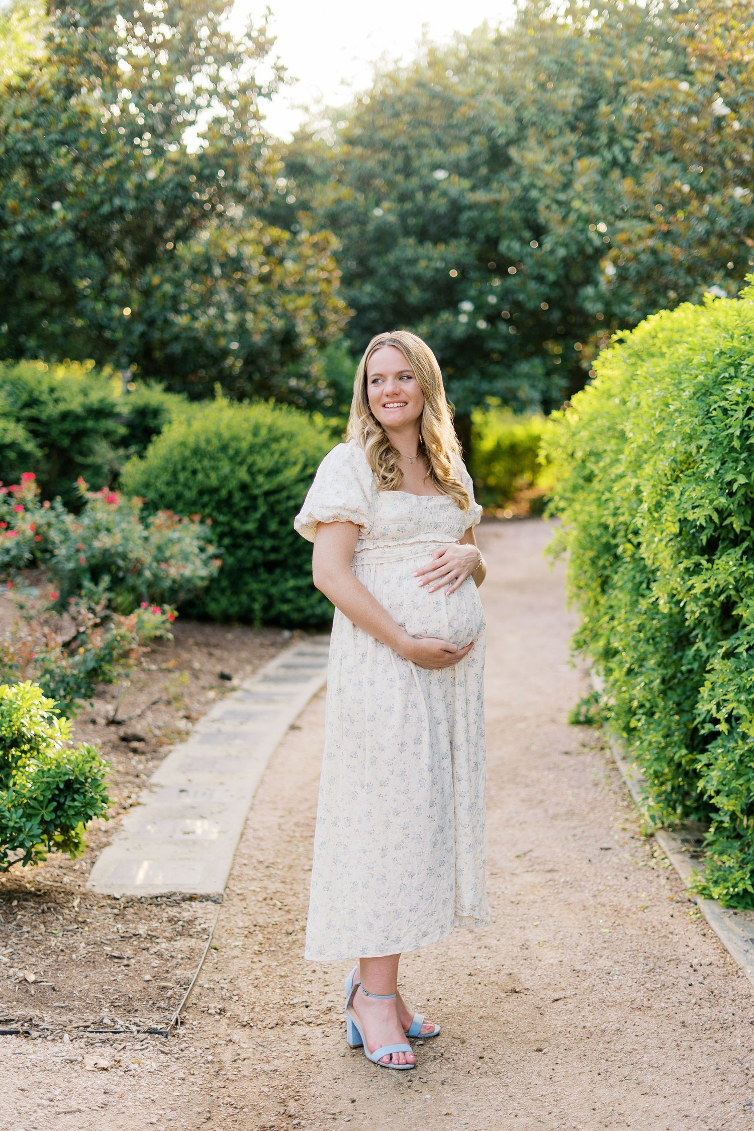 Woman in white floral dress poses in garden path during maternity photoshoot.