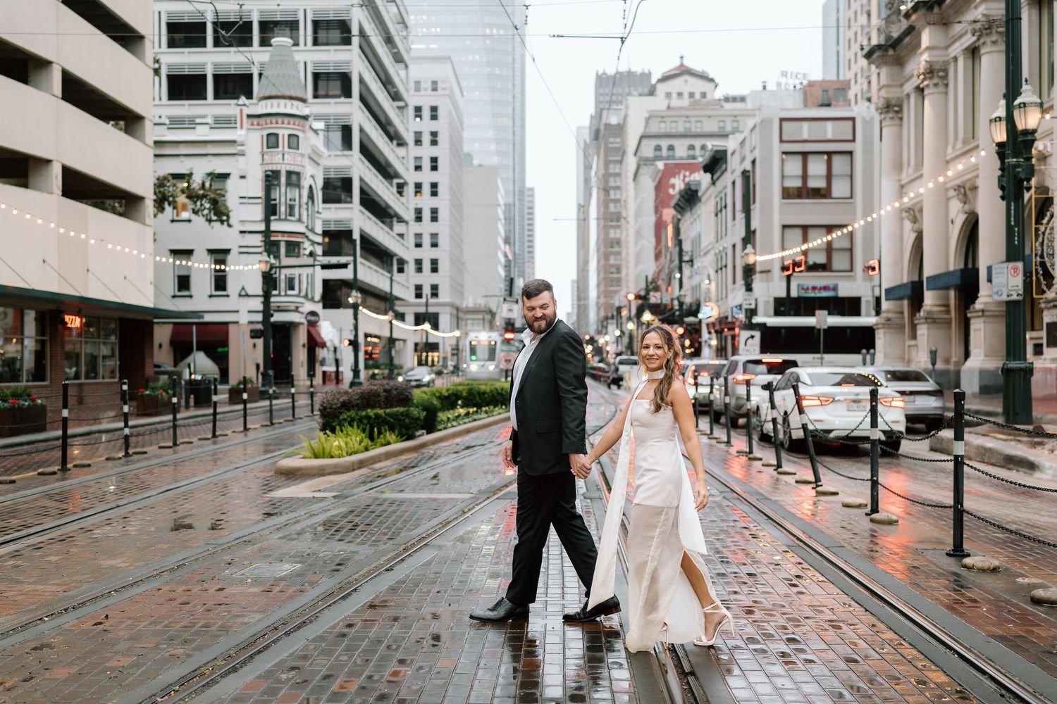 Couple walks through rainy city streets with streetcar tracks and high-rise buildings.