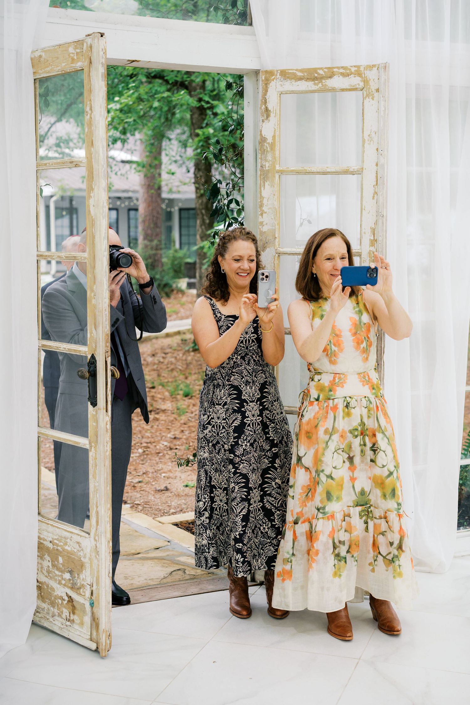 Two women in floral and patterned maxi dresses smile while taking photos at an elegant outdoor wedding venue.