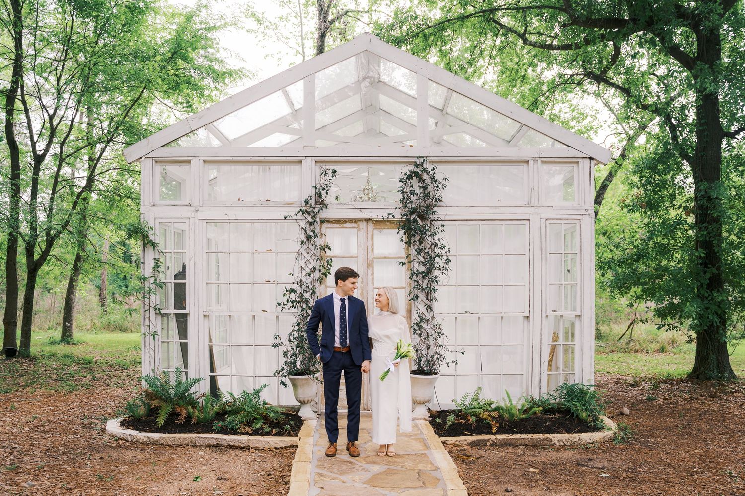 Bride and groom standing in front of a white greenhouse wedding venue surrounded by lush greenery and ivy.